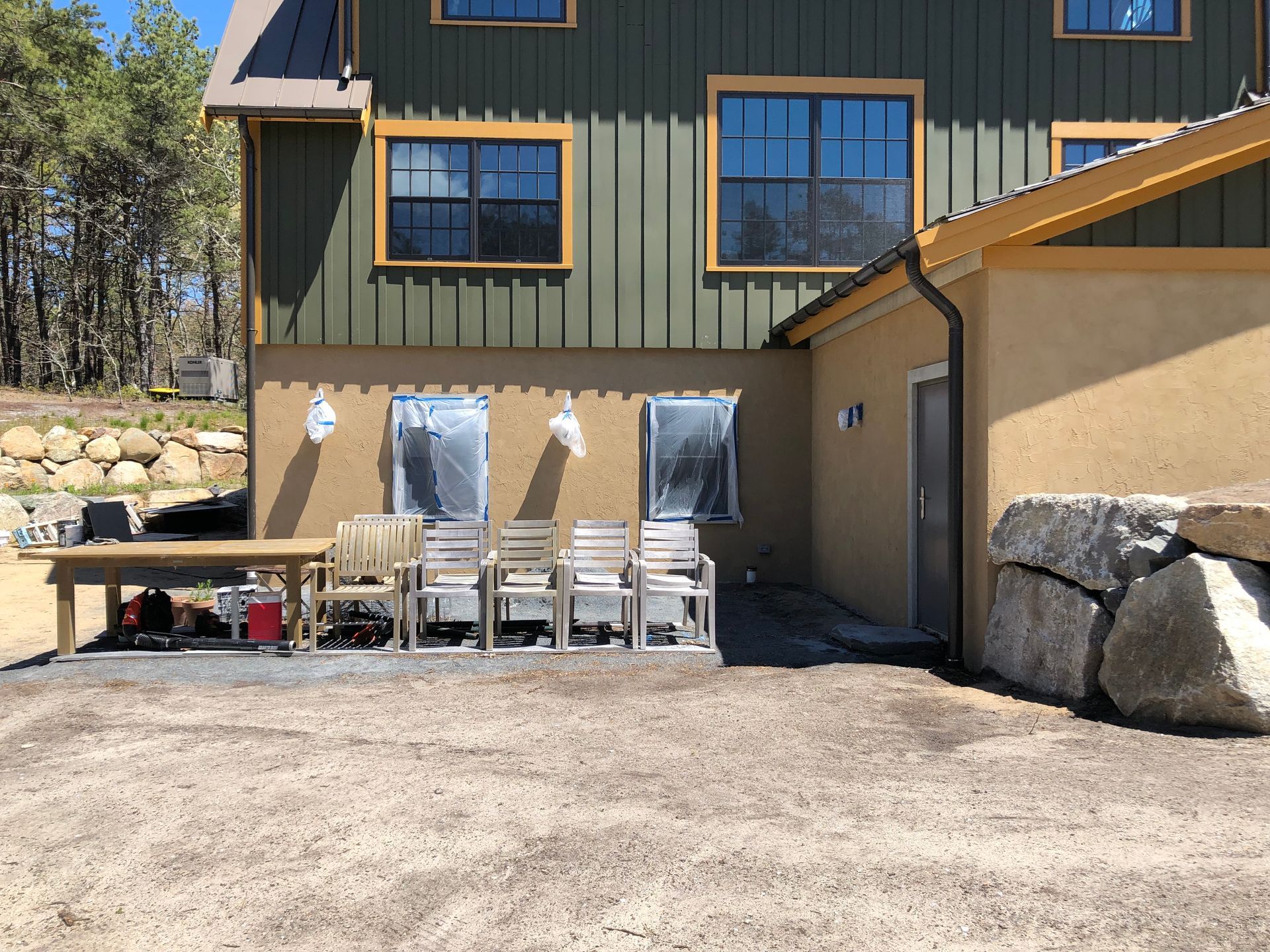 Outdoor patio beside a wood-sided house with folding chairs, a table, and large boulders along the wall