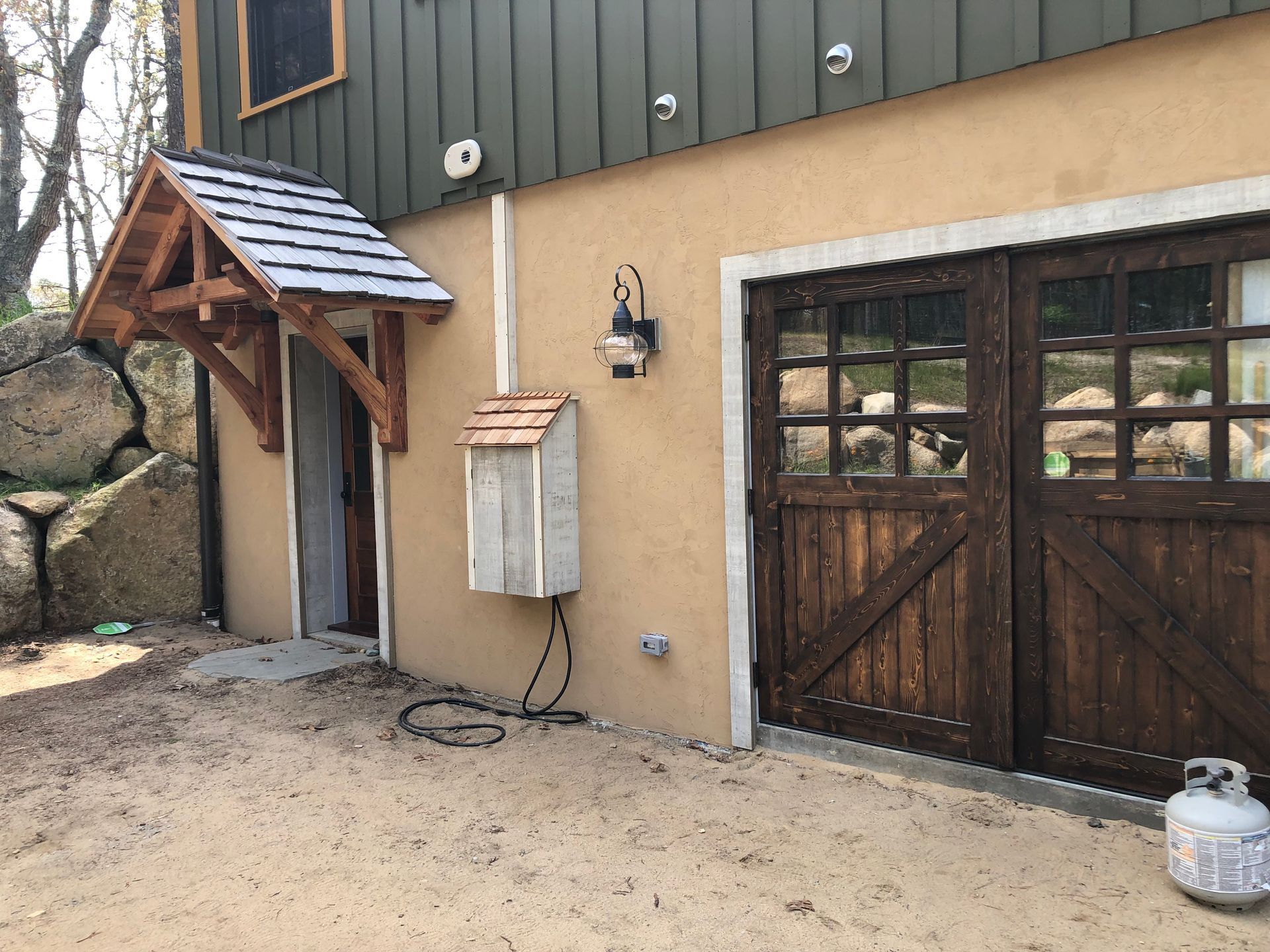 Rustic barn-style building with wooden double doors, side entrance canopy, and gas tank on sandy ground