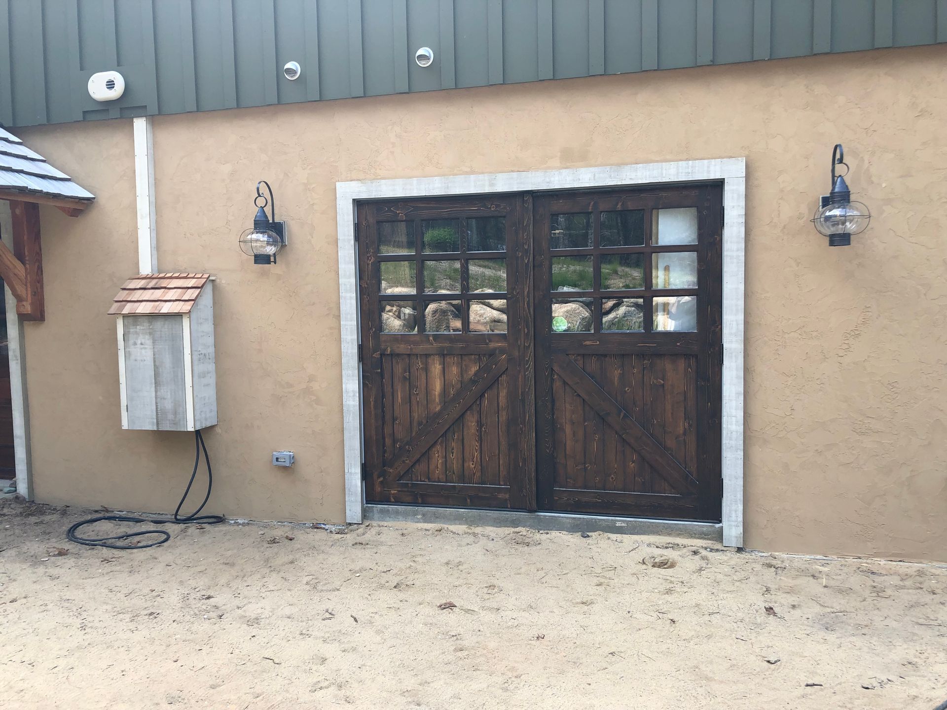 Rustic double wooden doors with windows on a tan exterior wall, flanked by two lanterns and a utility box.