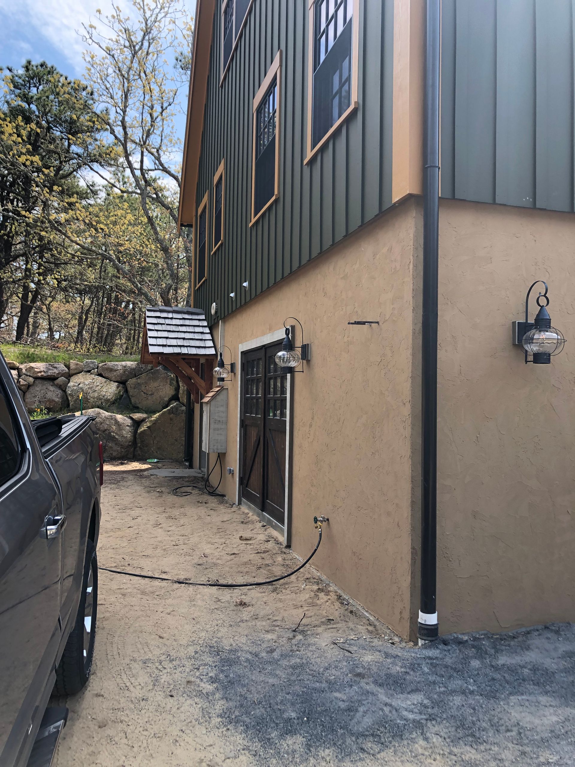 Side of a beige and dark green building with a narrow gravel walkway, parked vehicle, and trees nearby.