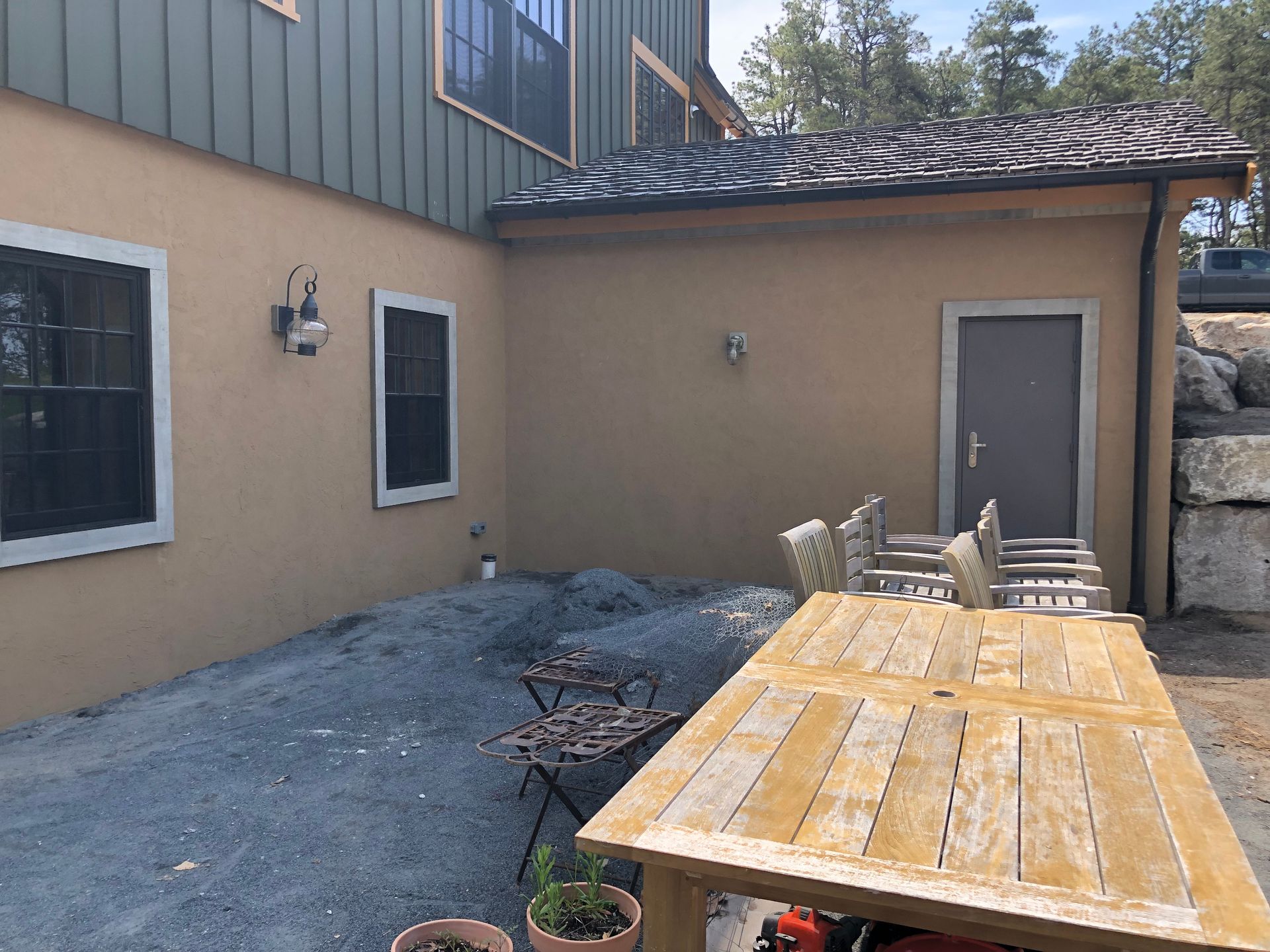 Patio with wooden table and chairs beside a tan house exterior and gravel yard