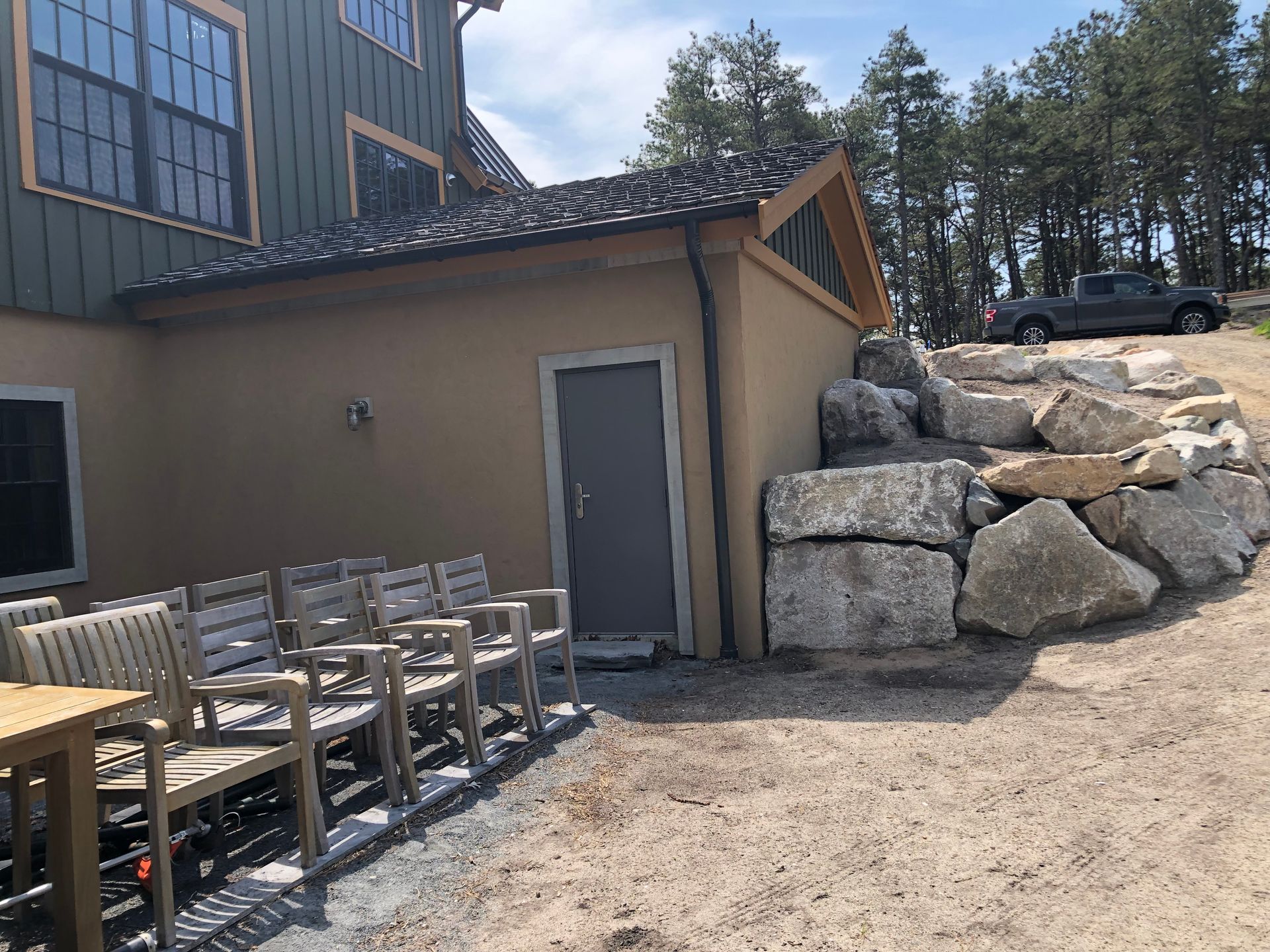 Outdoor patio beside a beige building with stacked chairs, a door, and large rocks under a wooded hillside.