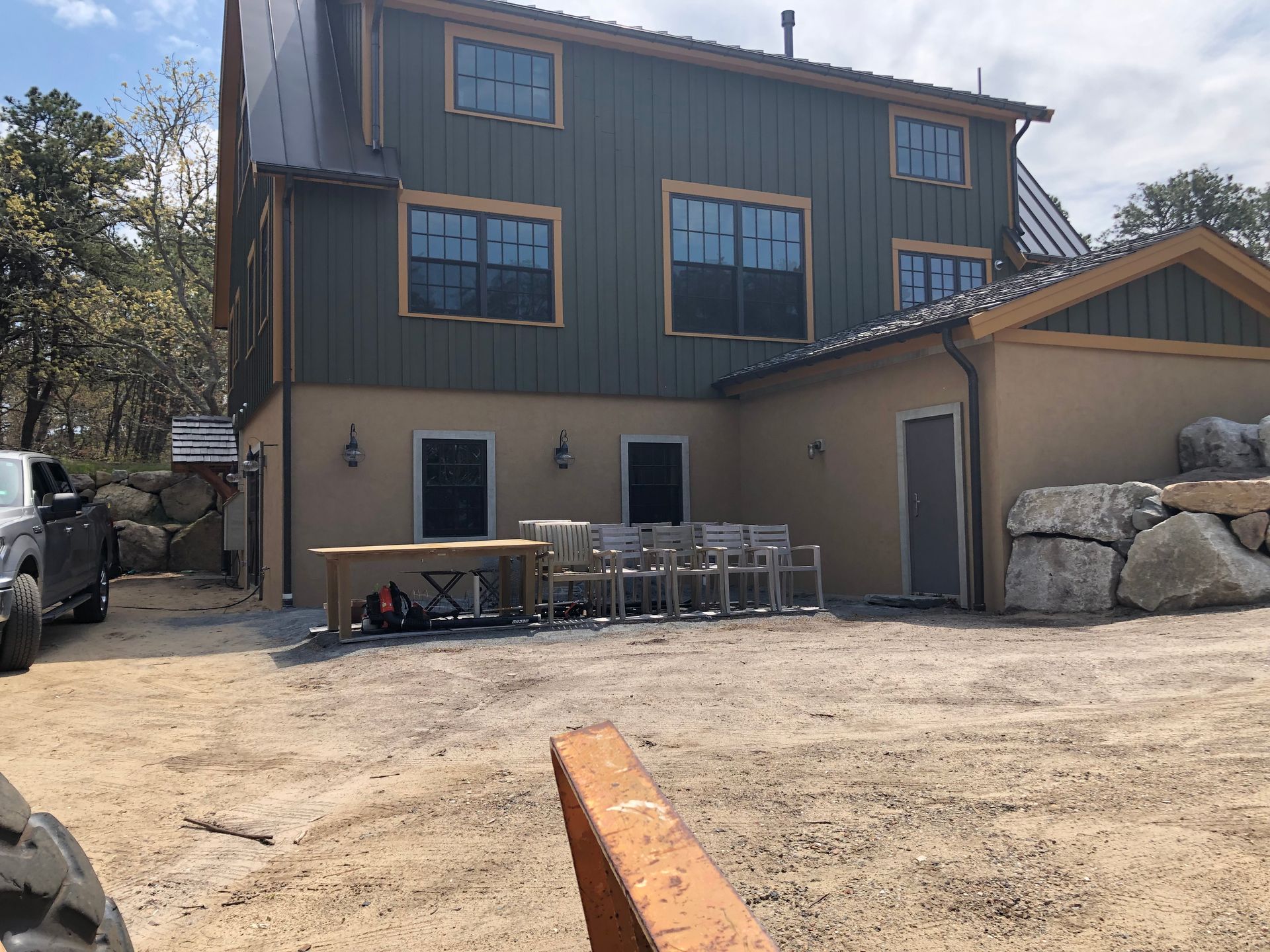 Two-story gray building with a small porch, stacked materials, and gravel lot in front