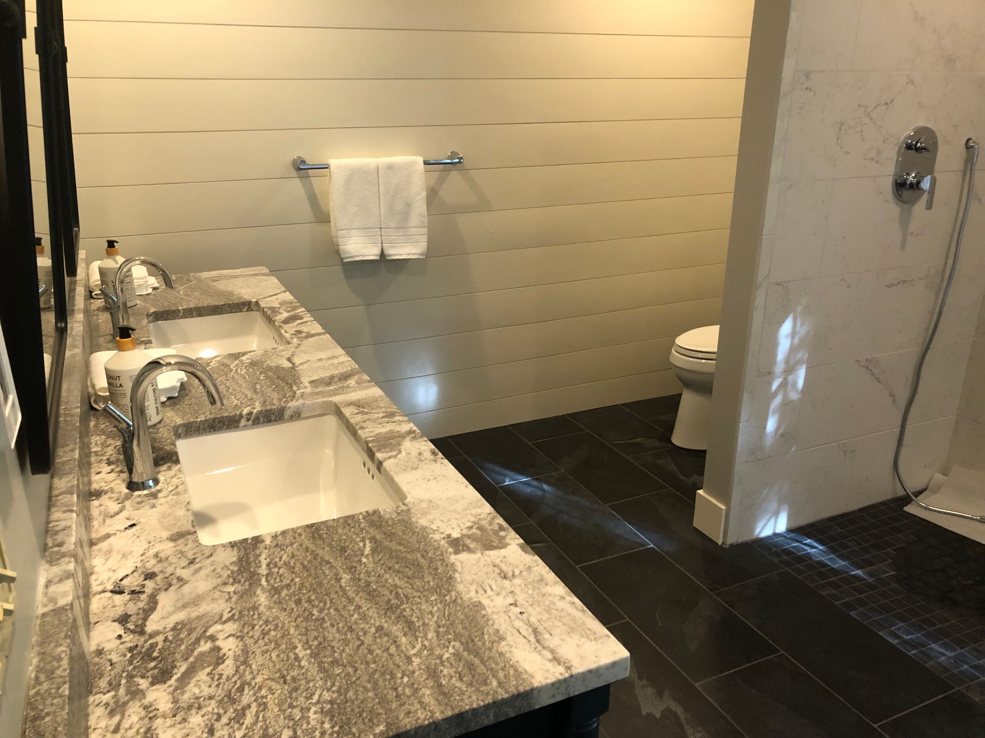 Modern bathroom with double sinks, marble countertop, dark tile floor, and toilet visible at right.