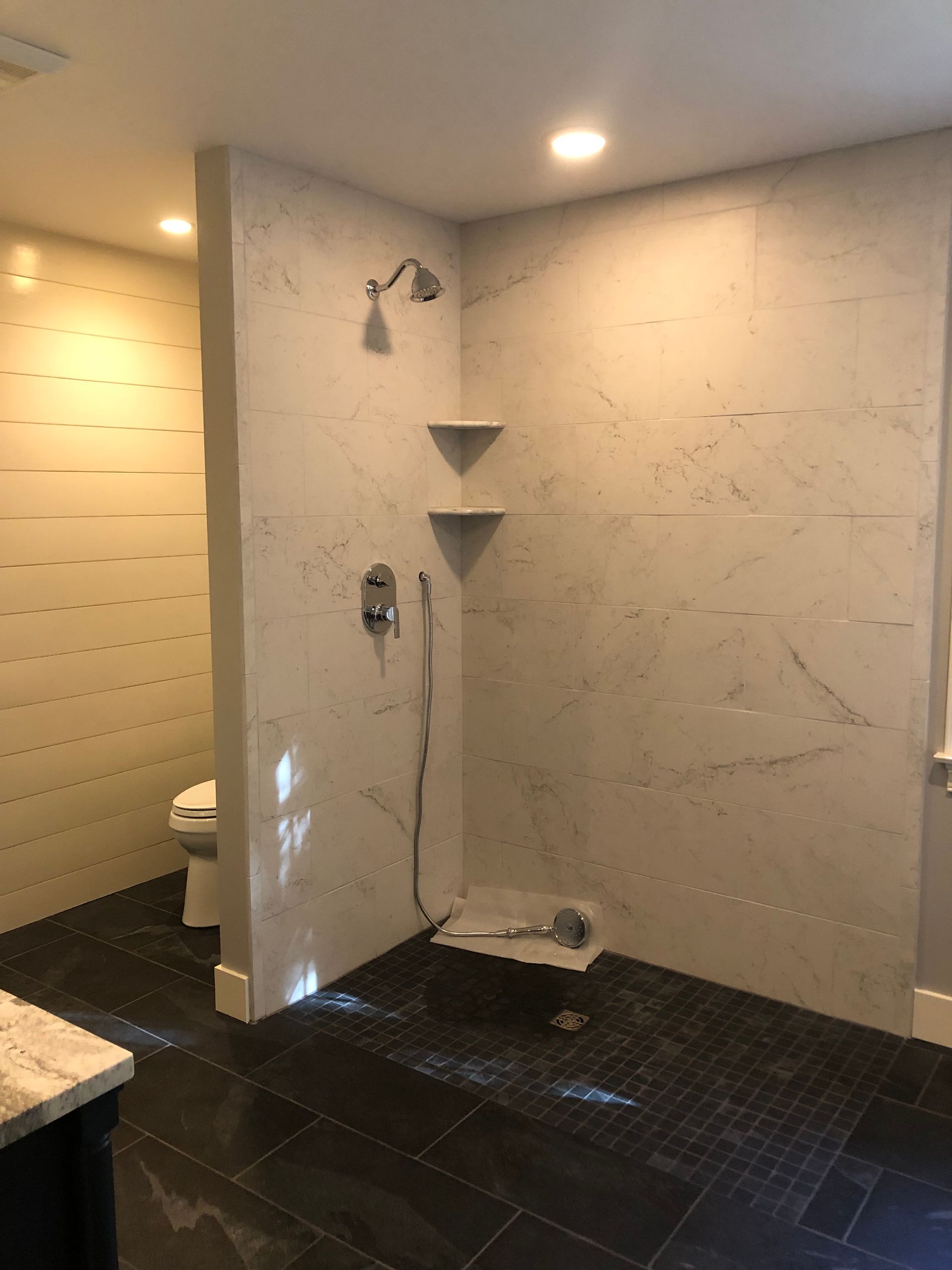 Tiled shower area with wall-mounted fixtures, corner shelves, and dark floor tiles in a bathroom