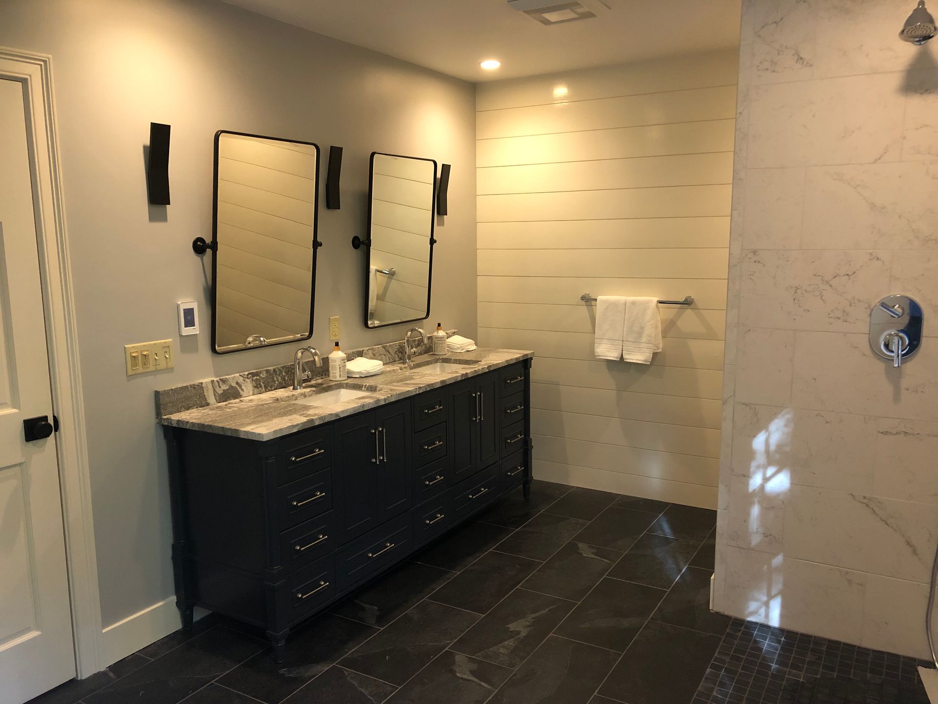 Modern bathroom with dark vanity, double mirrors, marble countertop, and beige tiled walls and floor