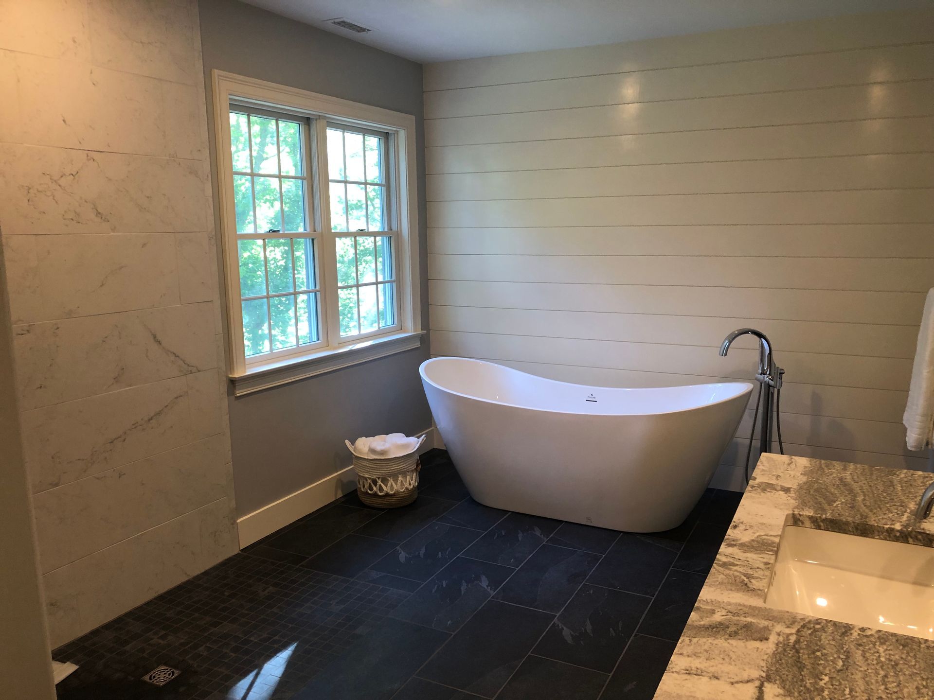Modern bathroom with a freestanding tub, window, dark tile floor, and marble vanity.