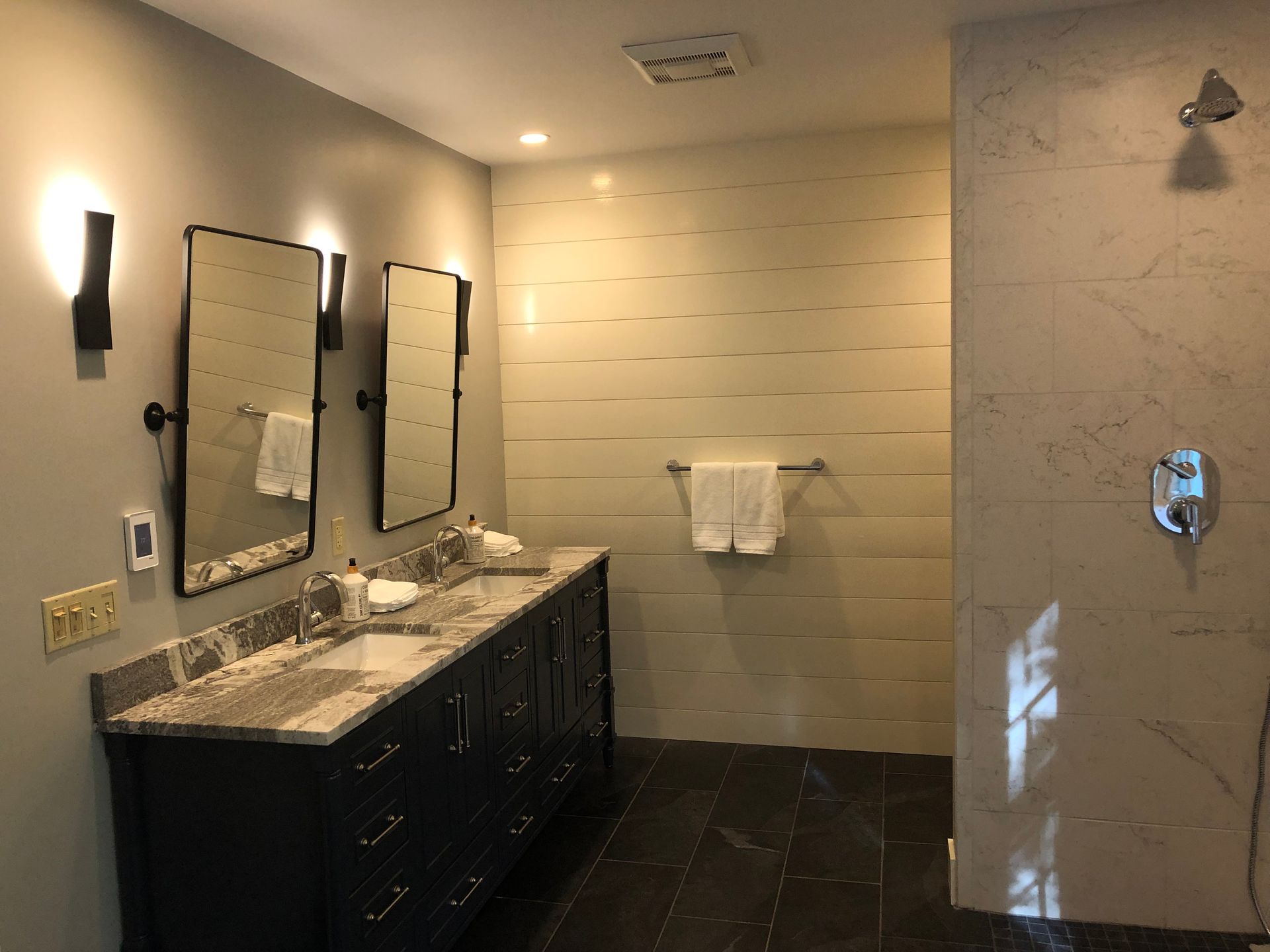 Modern bathroom with double vanity, mirrors, dark tile floor, and a glass shower.