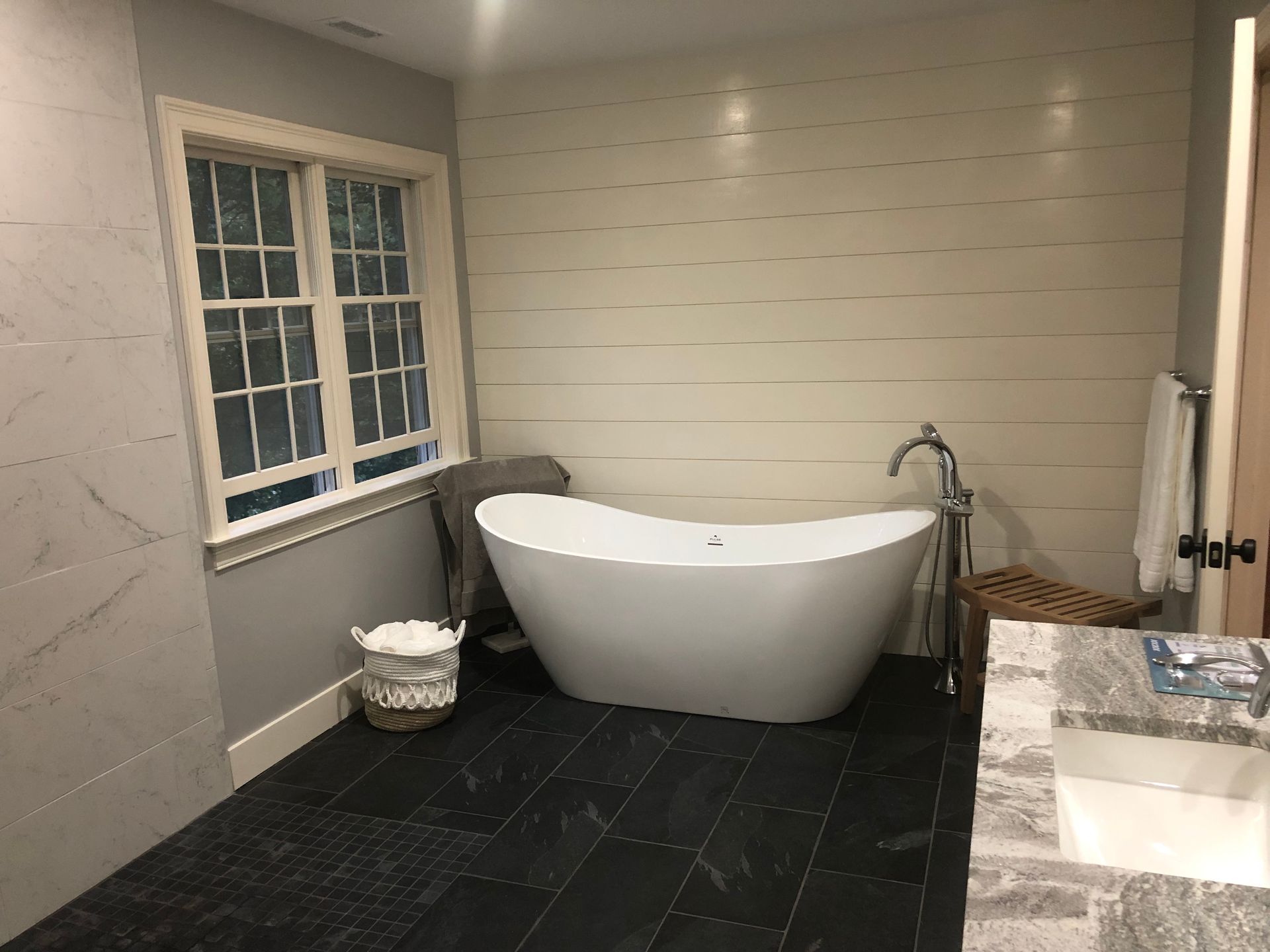 Modern bathroom with a freestanding white tub, window, dark tile floor, and vanity sink.