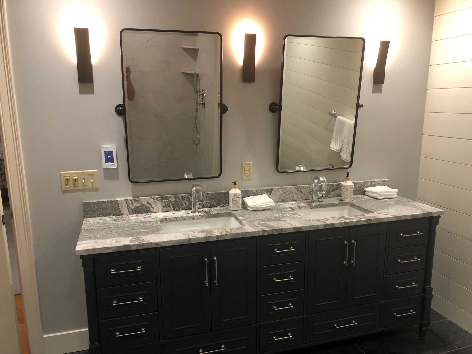 Double sink vanity with dark cabinets, two mirrors, wall sconces, and marble countertop in a bathroom