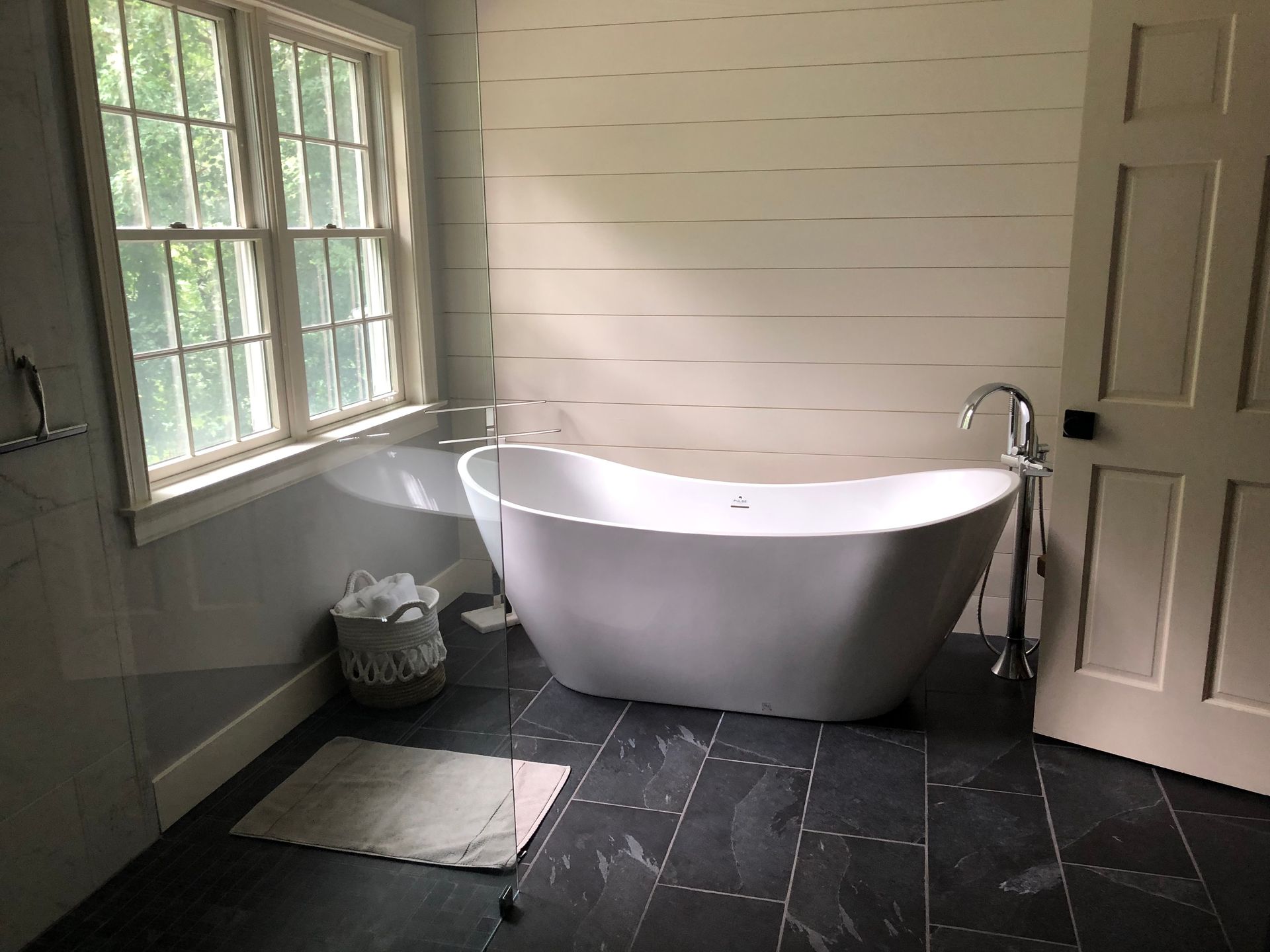 Bright bathroom with a freestanding white tub by a window, dark tile floor, and a small rug.