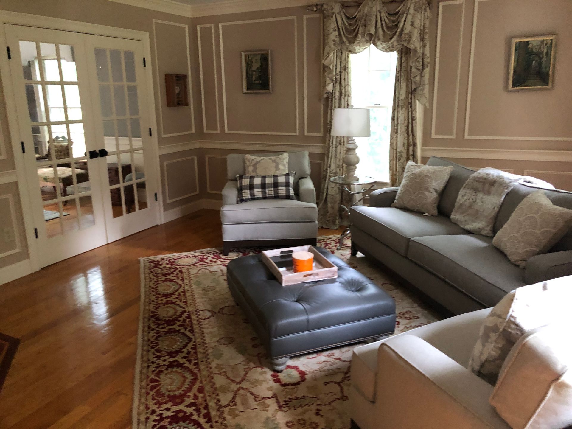 Elegant living room with gray sofa, ottoman, armchair, patterned rug, and French doors opening to bright light