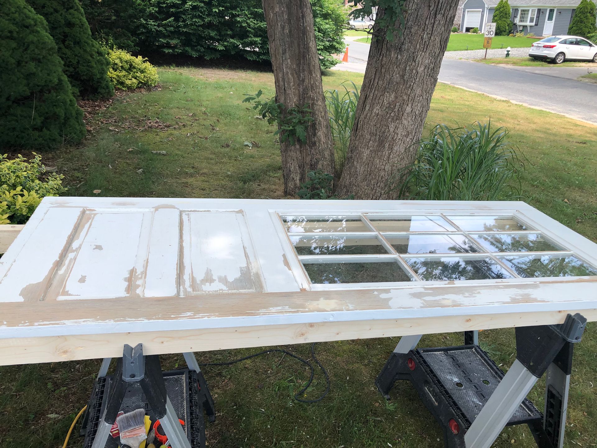 White door laid on sawhorses outdoors, with a windowpane section and paint along the edges.