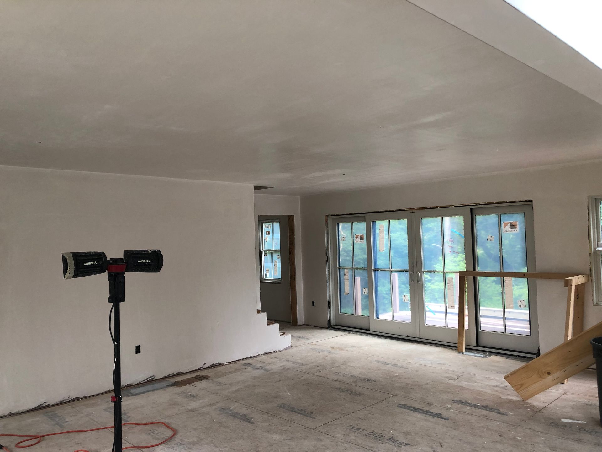 Unfinished living room with bare walls, exposed floors, large windows, and a work light on a stand