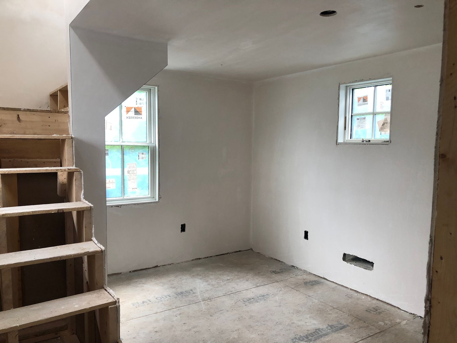 Empty unfinished room with bare drywall, two small windows, and stairs on the left.