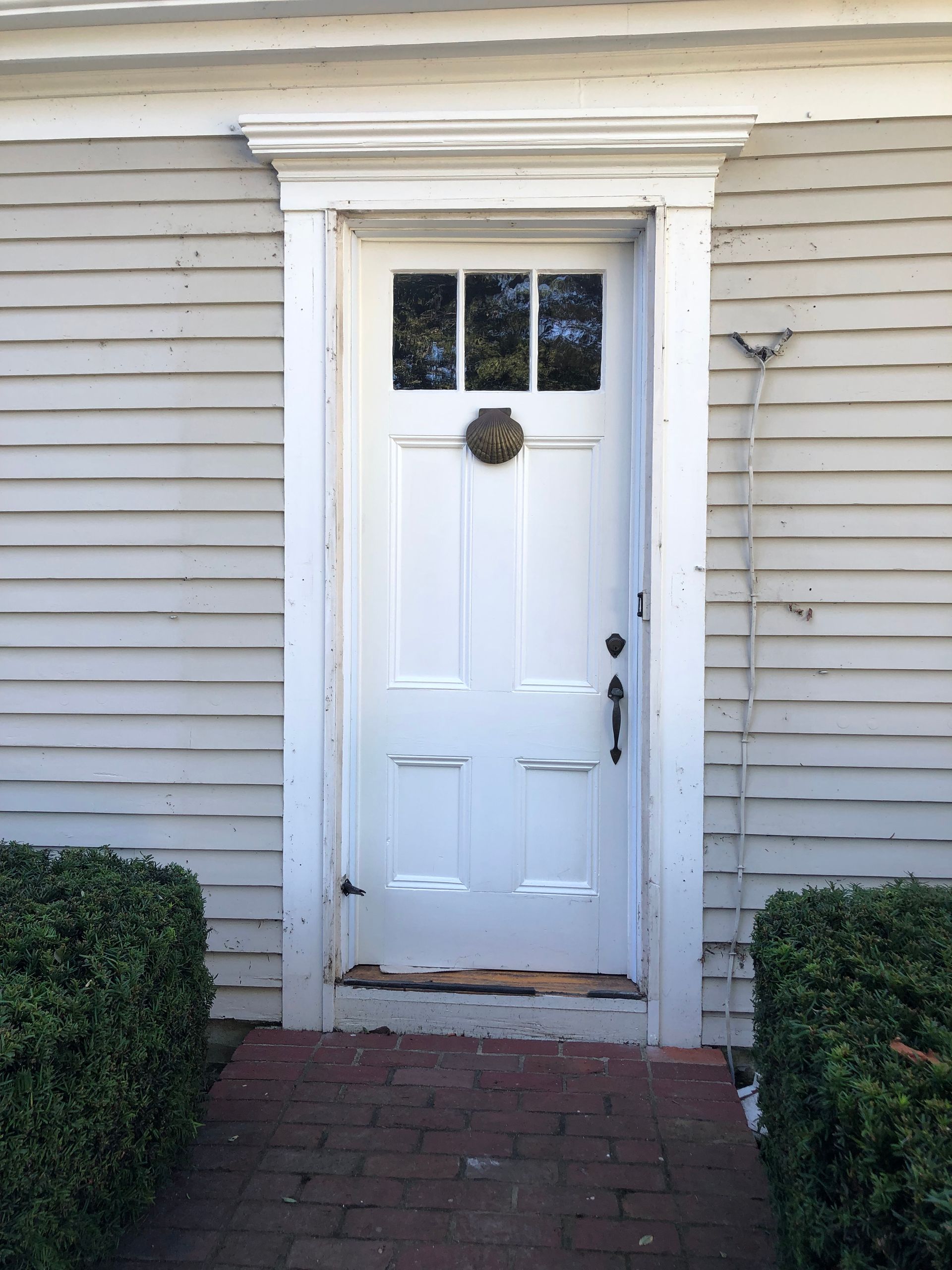 White front door with small window panes, brick walkway, and bushes beside gray siding.