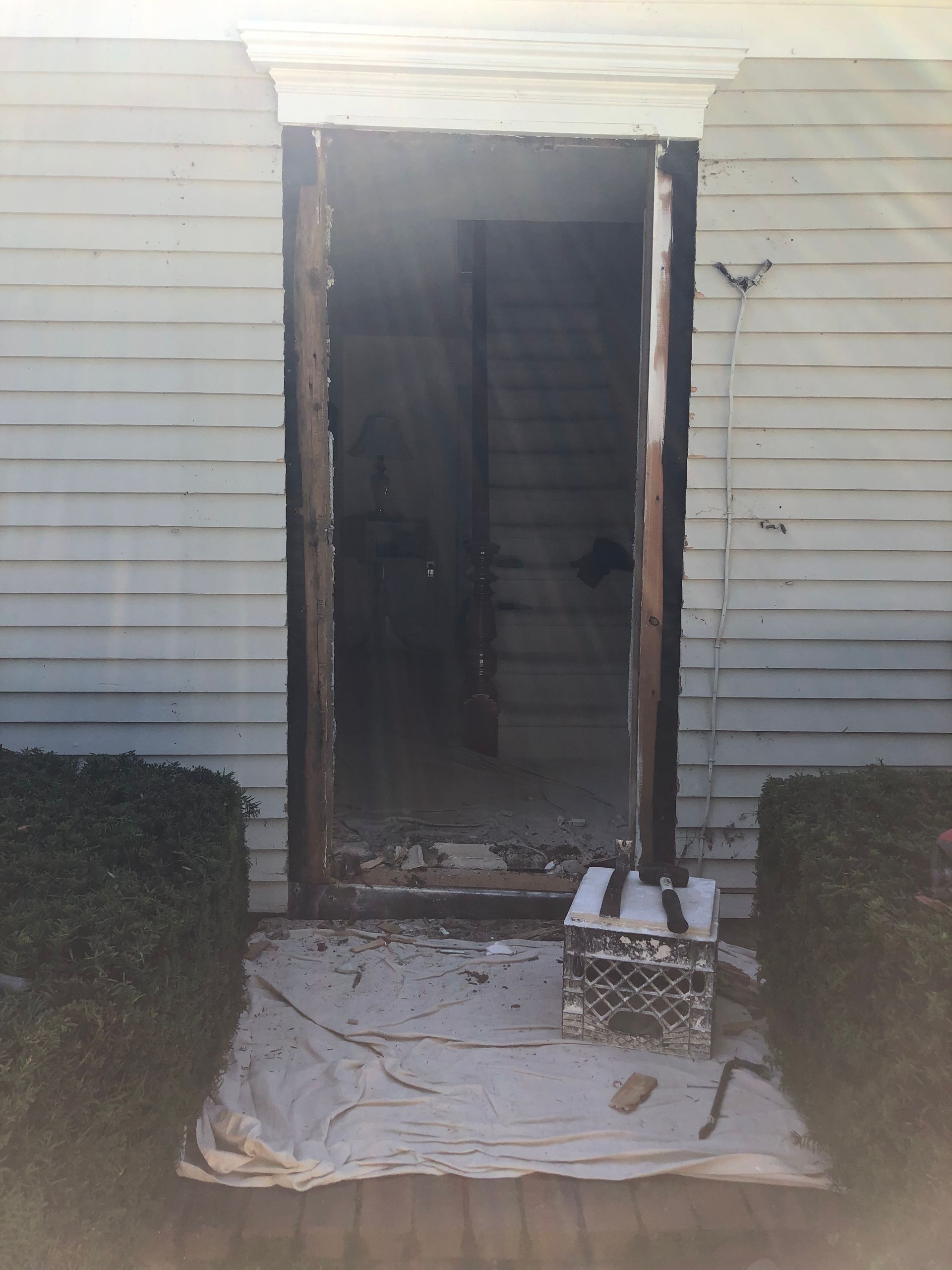 Front door of a house with damaged trim, an open doorway, and debris on a white drop cloth.