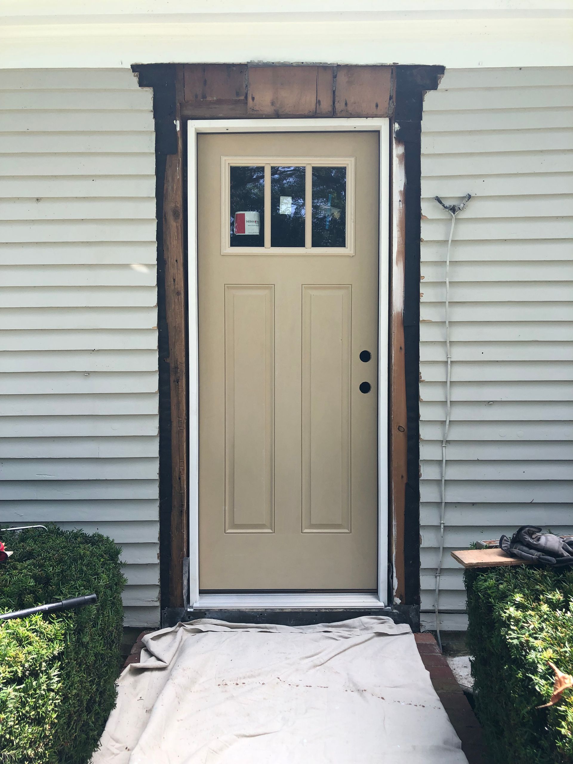 Beige front door with small window in white siding, framed by dark trim and a concrete walkway