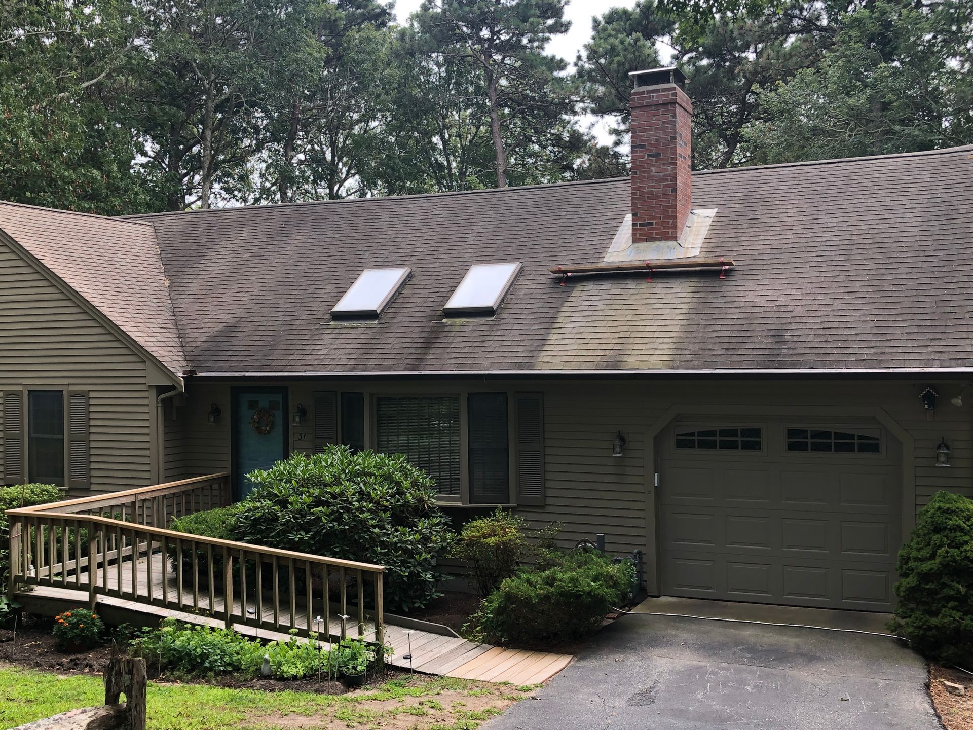 Suburban house with dark shingle roof, two skylights, brick chimney, garage, and front ramp surrounded by shrubs