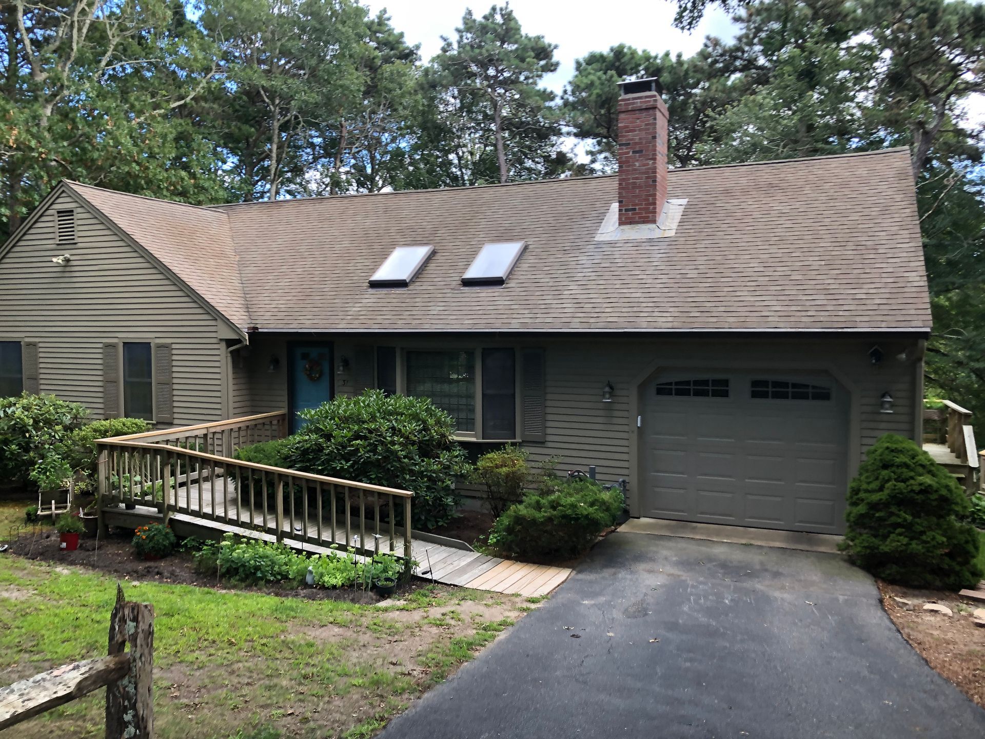 Single-story house with gray roof, garage, ramp, and driveway surrounded by trees.