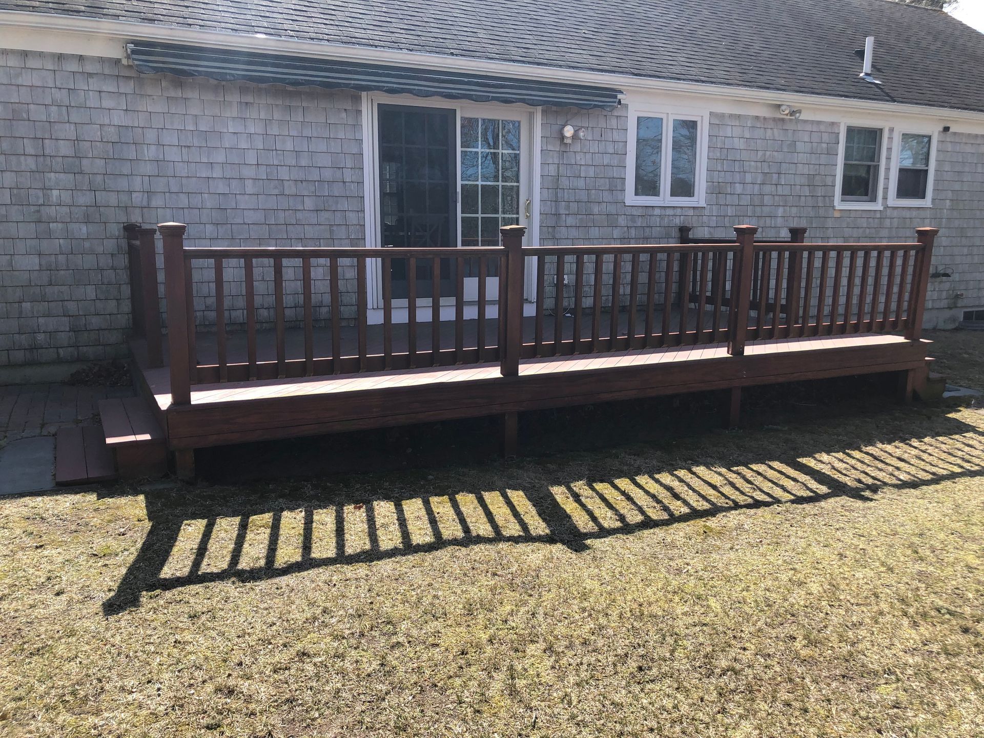 Back deck with brown railing attached to a gray house, casting striped shadows on the lawn.