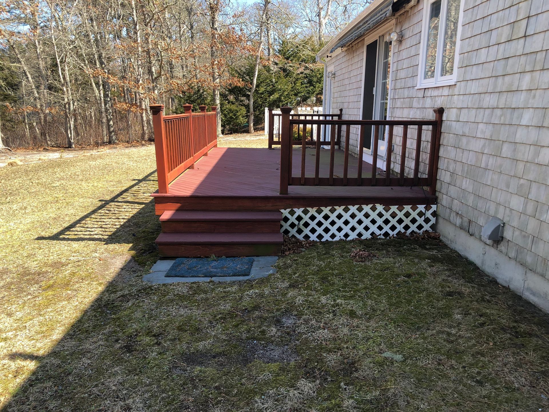 Small wooden porch with steps, red railing, and lattice skirting beside a stone house in a yard.