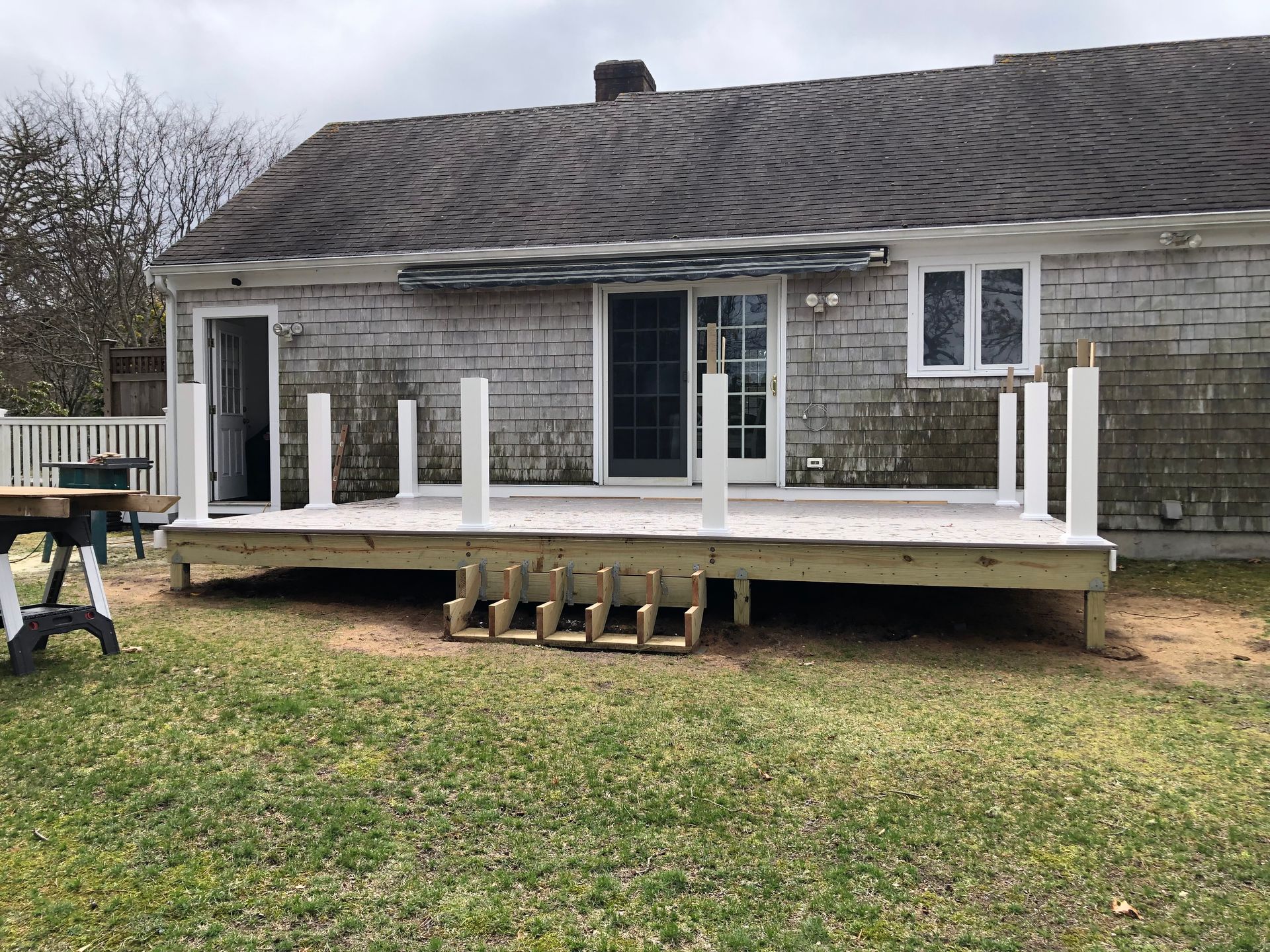 Raised wooden deck under construction behind a weathered house, with white posts and ladders beneath it
