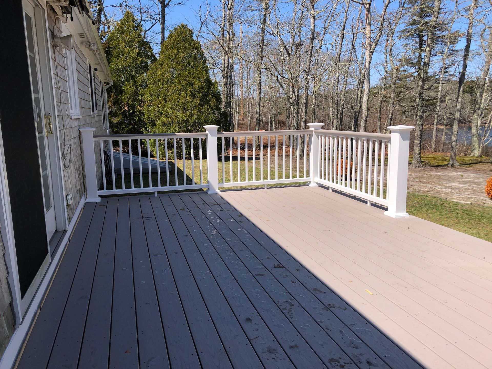 Wooden deck with white railing beside a house, overlooking trees and a driveway.