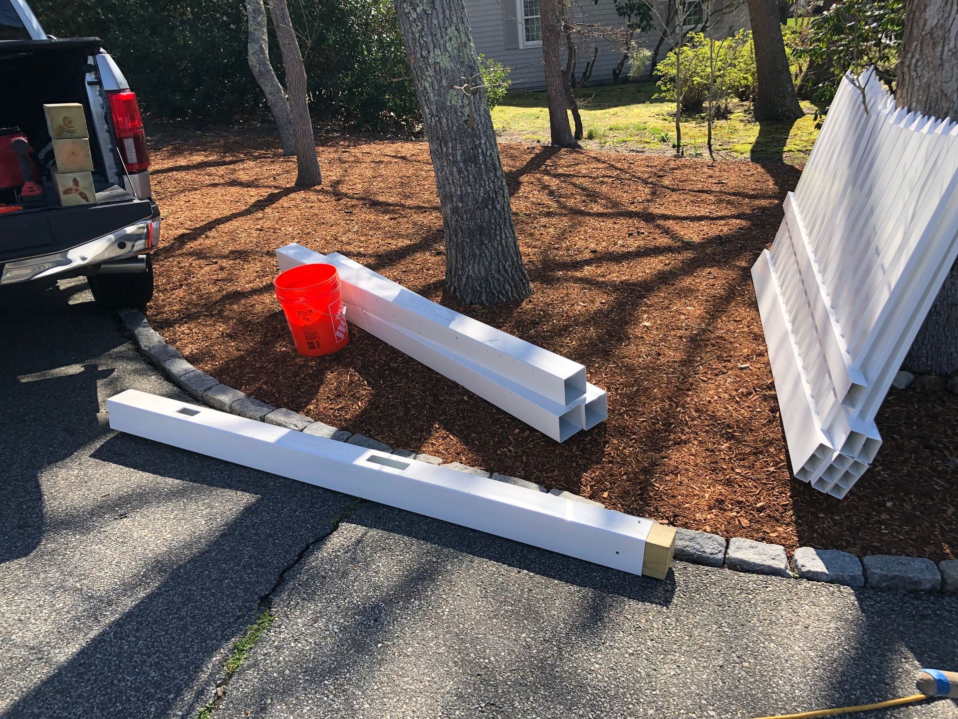 Broken white vinyl fence pieces beside a curb, with a red bucket and a black truck nearby.