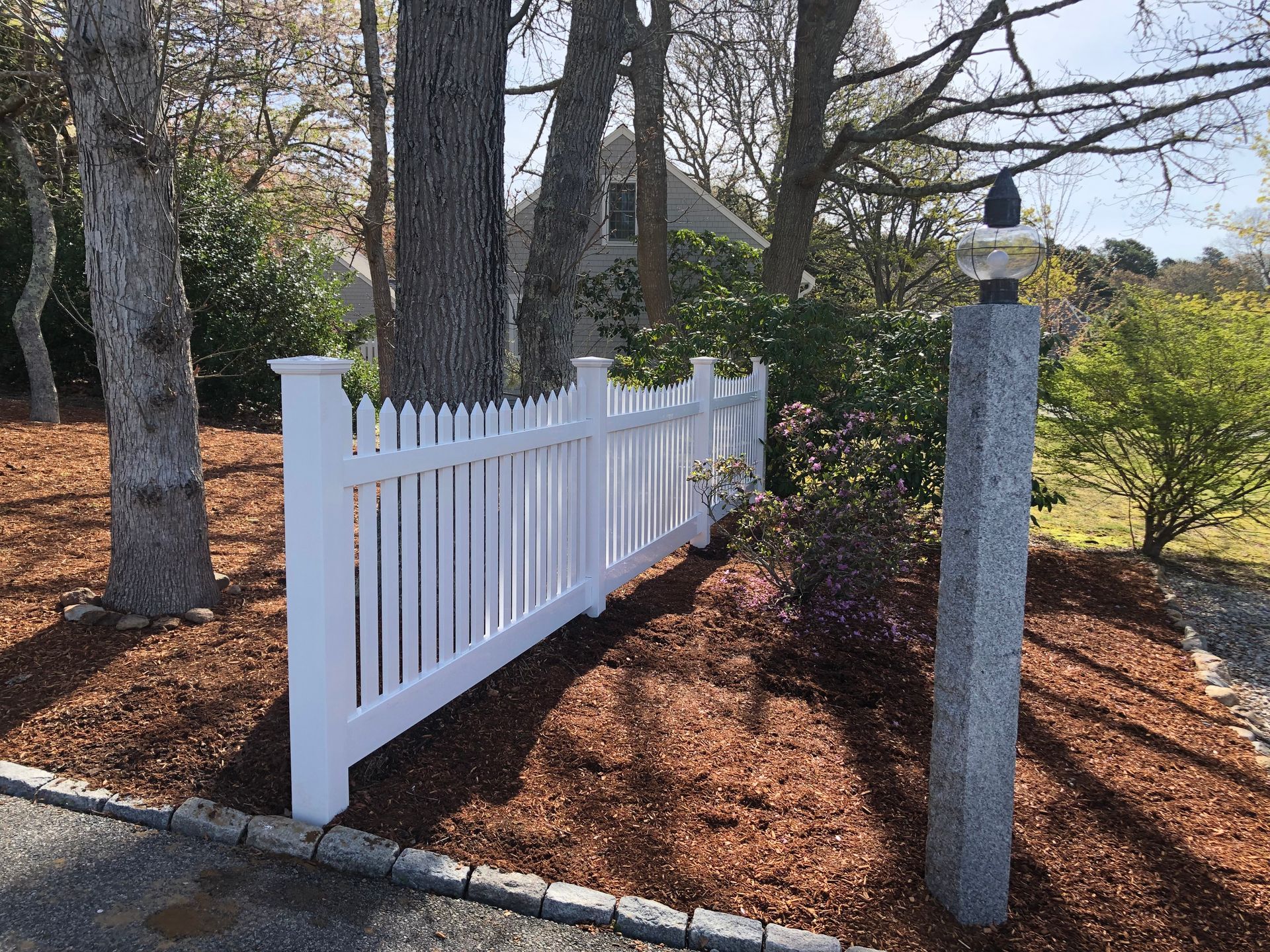White picket fence beside trees and mulch in a landscaped yard, with a stone post in front.