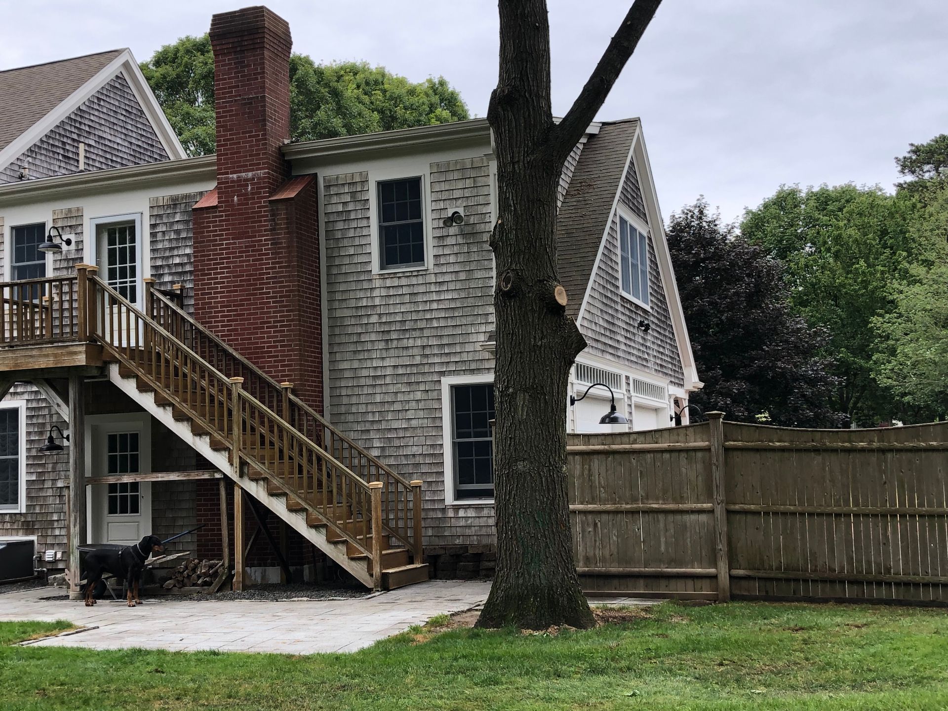 Exterior of a house with an elevated wooden staircase, brick chimney, and fenced yard under trees