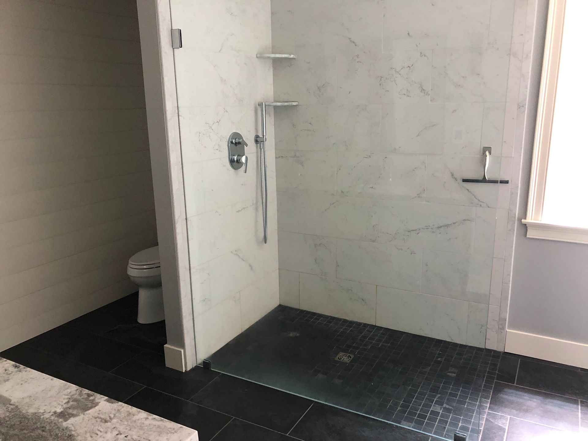 Modern shower with black tile floor, white tiled walls, grab bar, and a visible toilet in an adjacent space