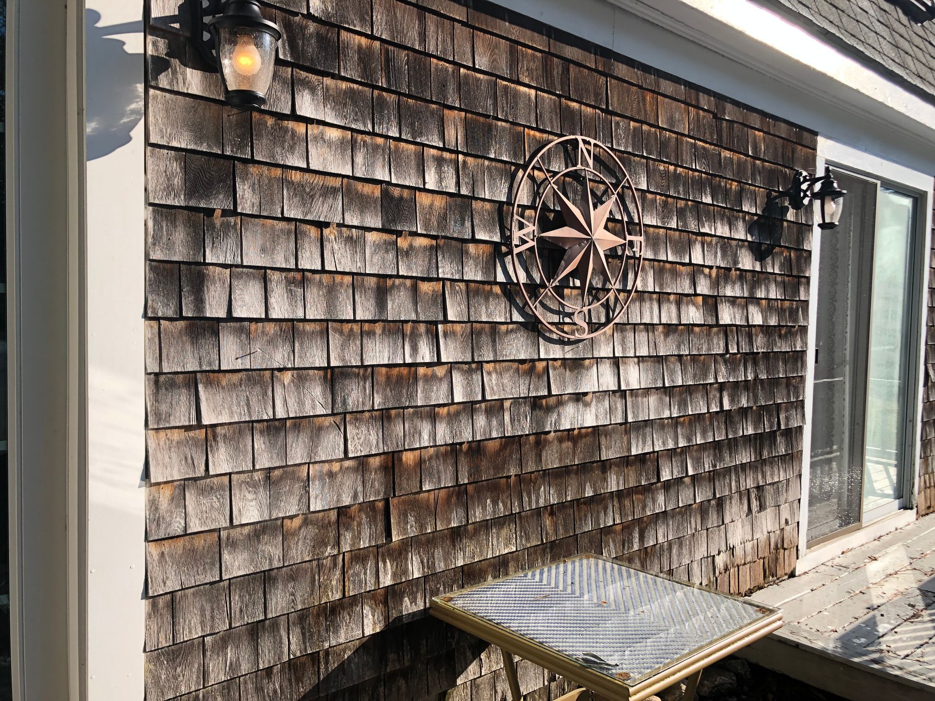 Weathered shingle exterior wall with a nautical wheel decor, lanterns, and a small bench by a glass door