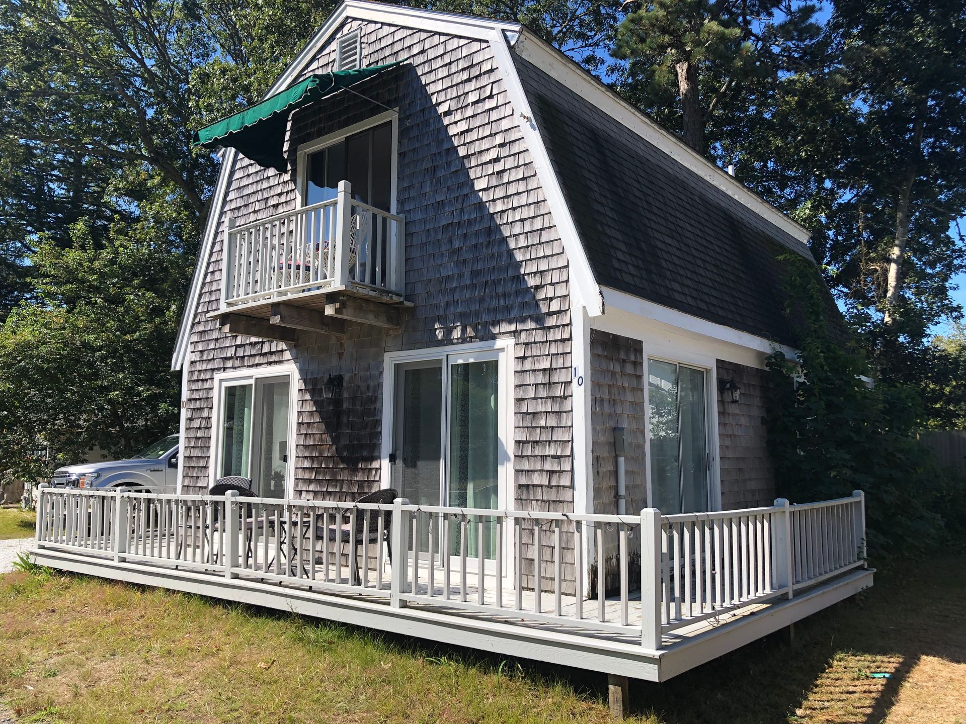 Small gray cottage with white trim and a wraparound porch in a wooded yard
