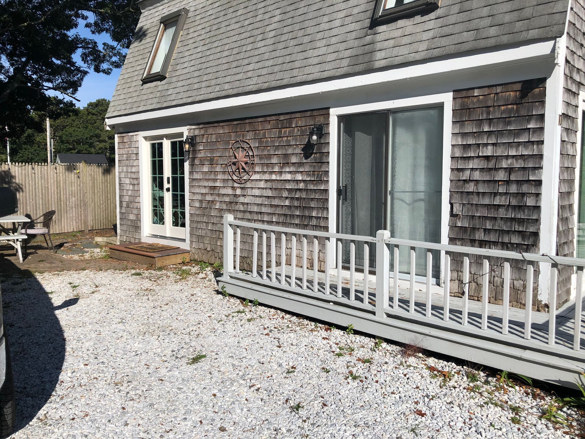 Weathered shingle house with sliding glass doors, white railing, and a gravel yard in front