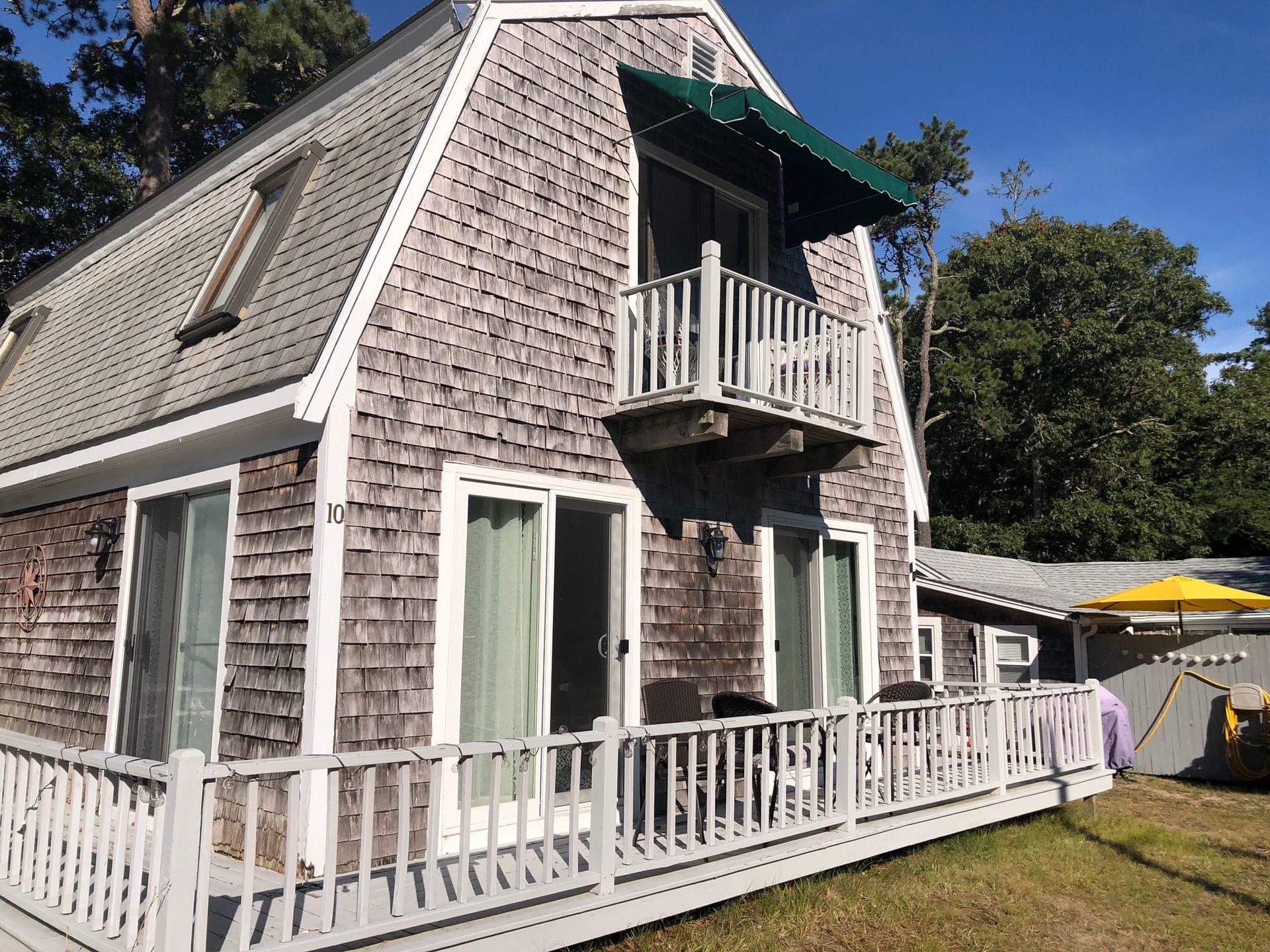 Cape-style gray shingled house with white trim and balconies, fenced yard, and a patio umbrella on the right.