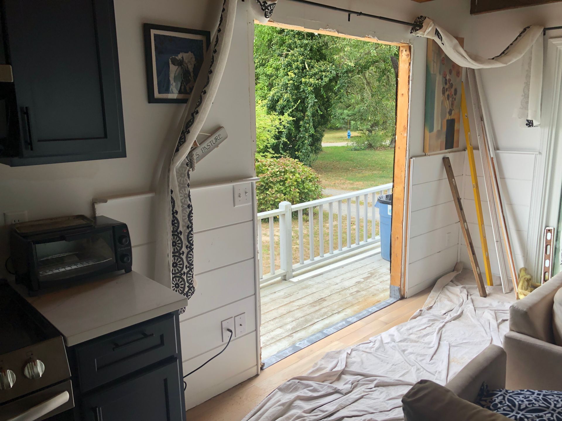 Small room with a broken wall opening to a deck and trees outside, white tarp on the floor.