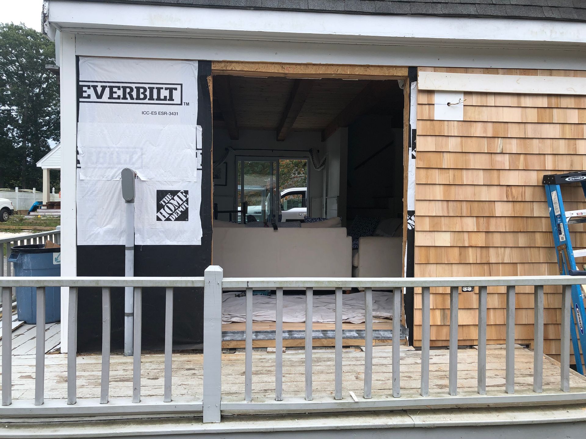Partially built house exterior with open doorway, white siding, wood shingles, and a safety railing in front