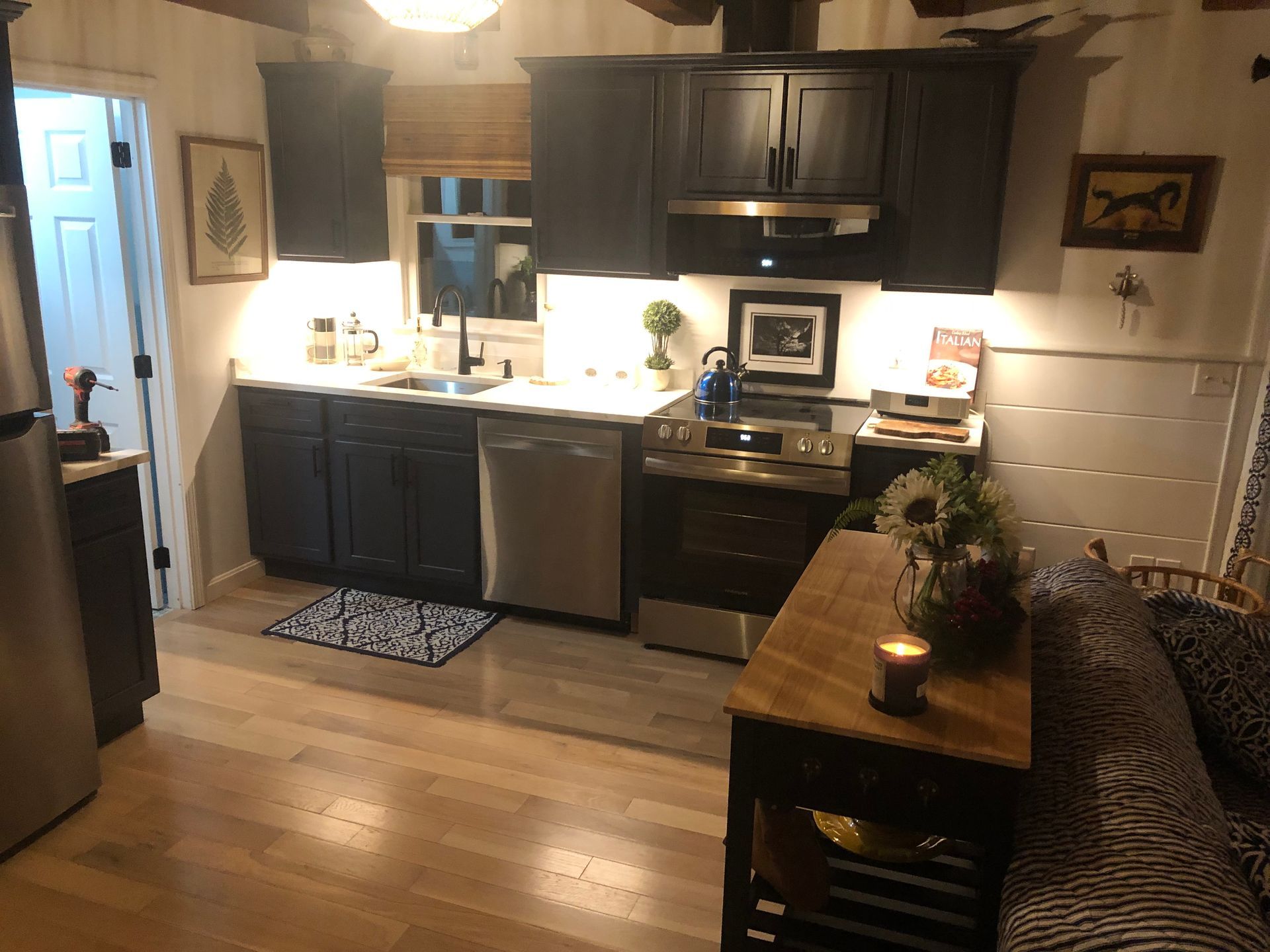 Warm kitchen with dark cabinets, stainless appliances, wood floors, and a small table beside a sofa.