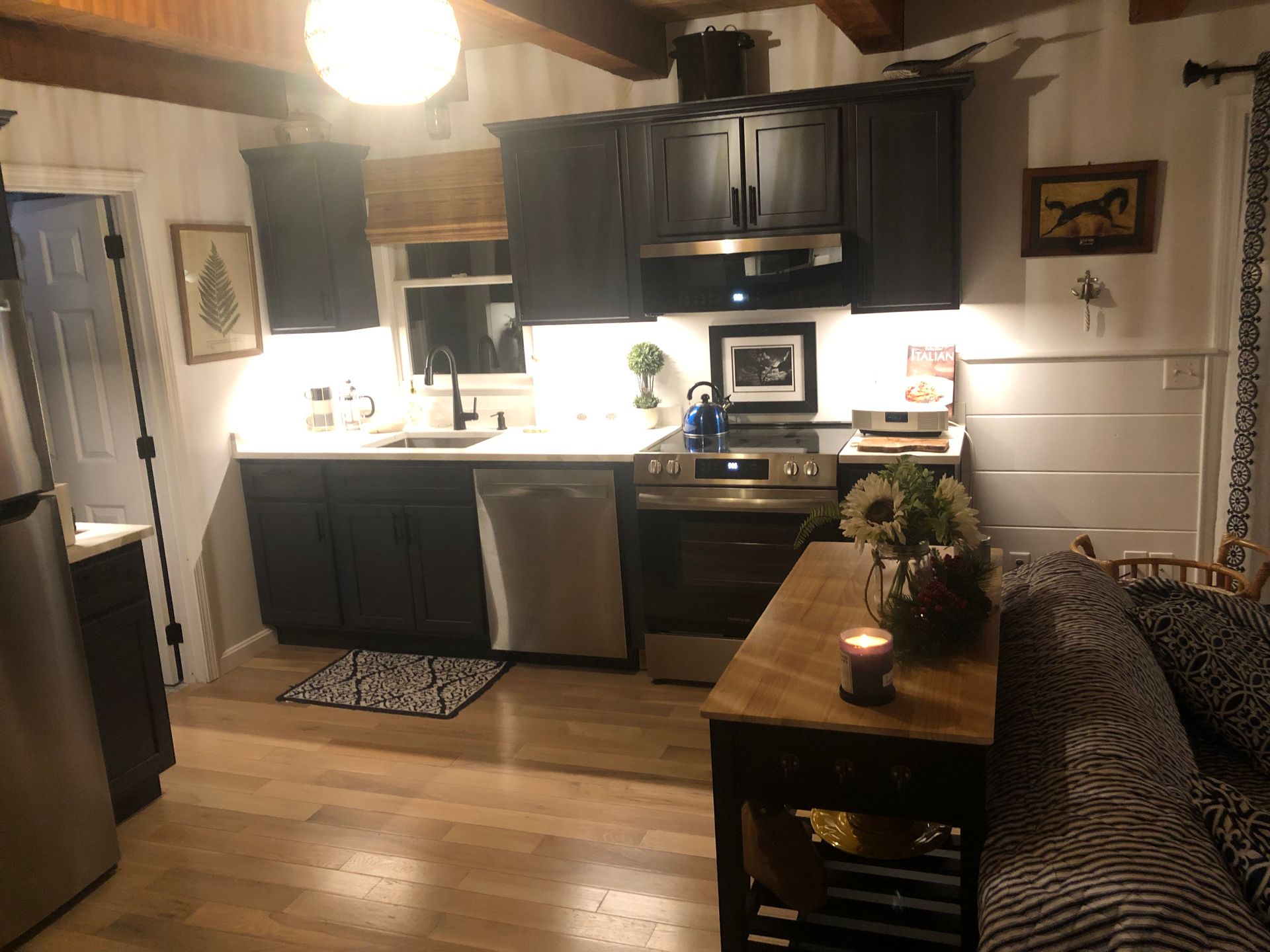 Cozy kitchen with dark cabinets, white counters, stove, and a small wooden table beside a patterned sofa.