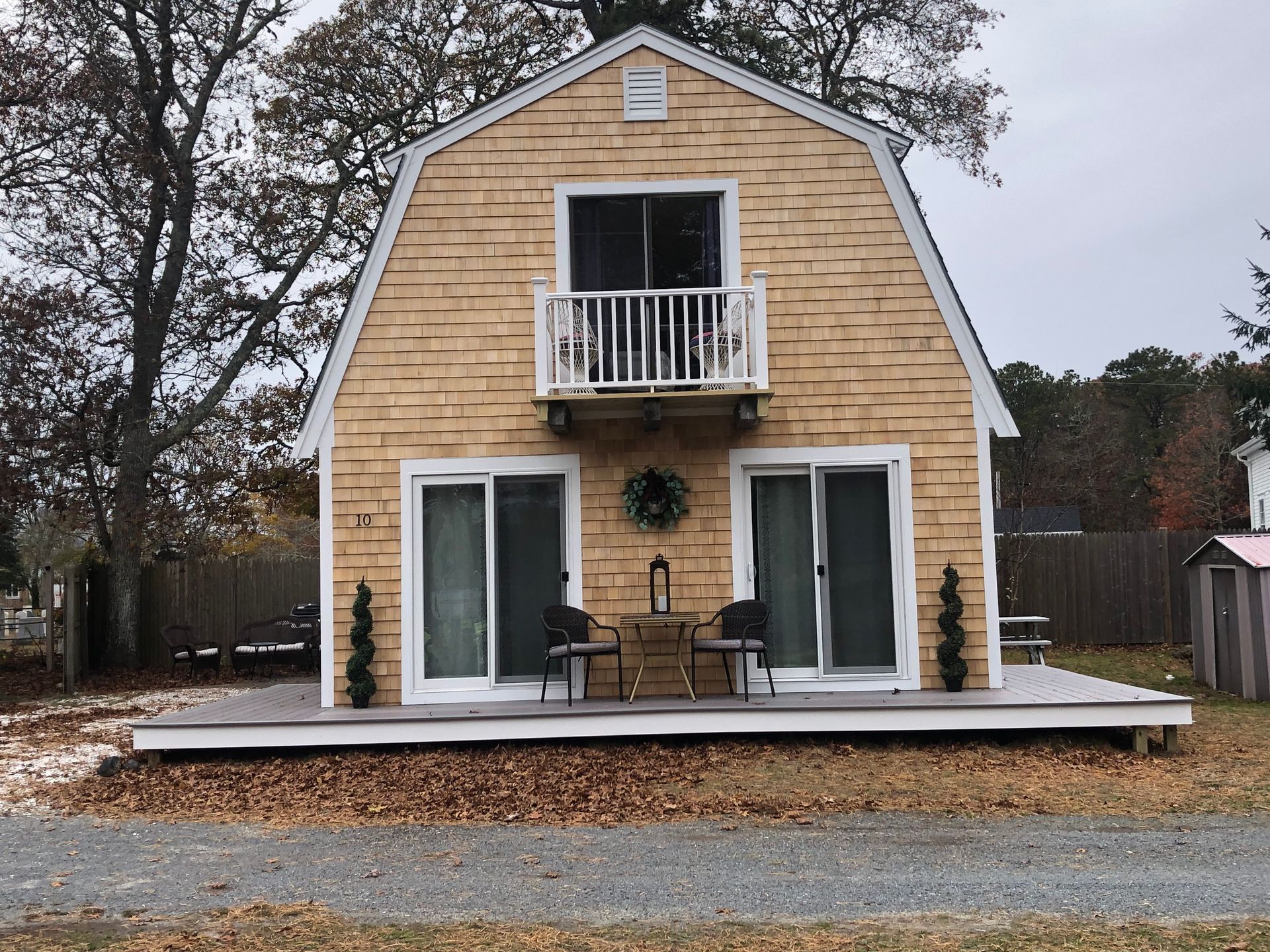 Small tan barn-style house with white trim and a front porch, centered on a gravel lot amid autumn trees