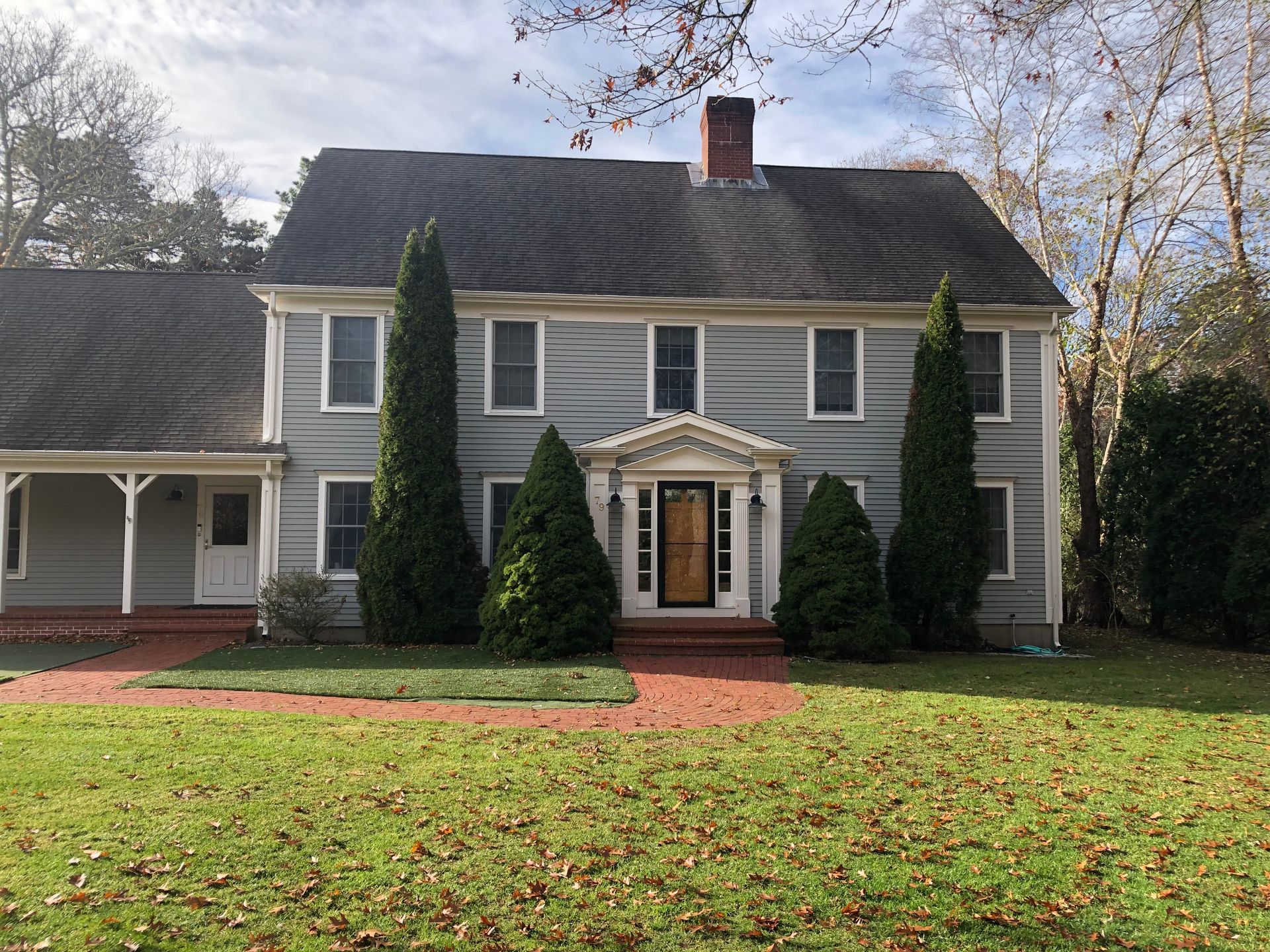 Gray two-story house with white trim, brick walkway, and tall evergreen trees in front lawn