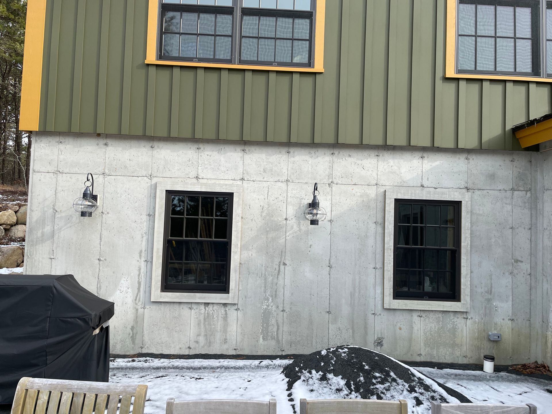 Exterior of a house with gray concrete lower wall, olive siding above, and two black-framed windows in winter snow