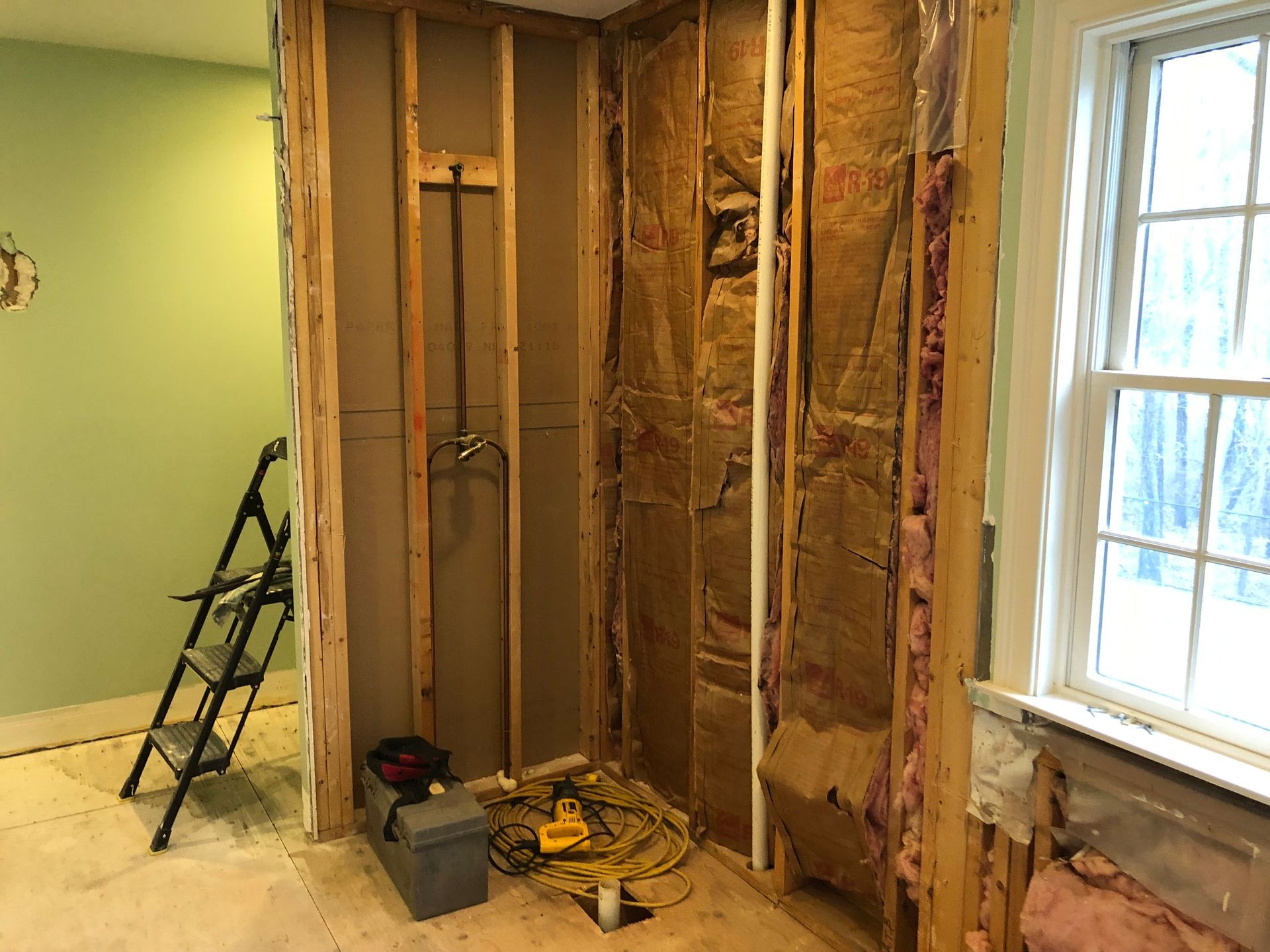 Partially gutted room with exposed studs, insulation, tools, and a ladder by a window