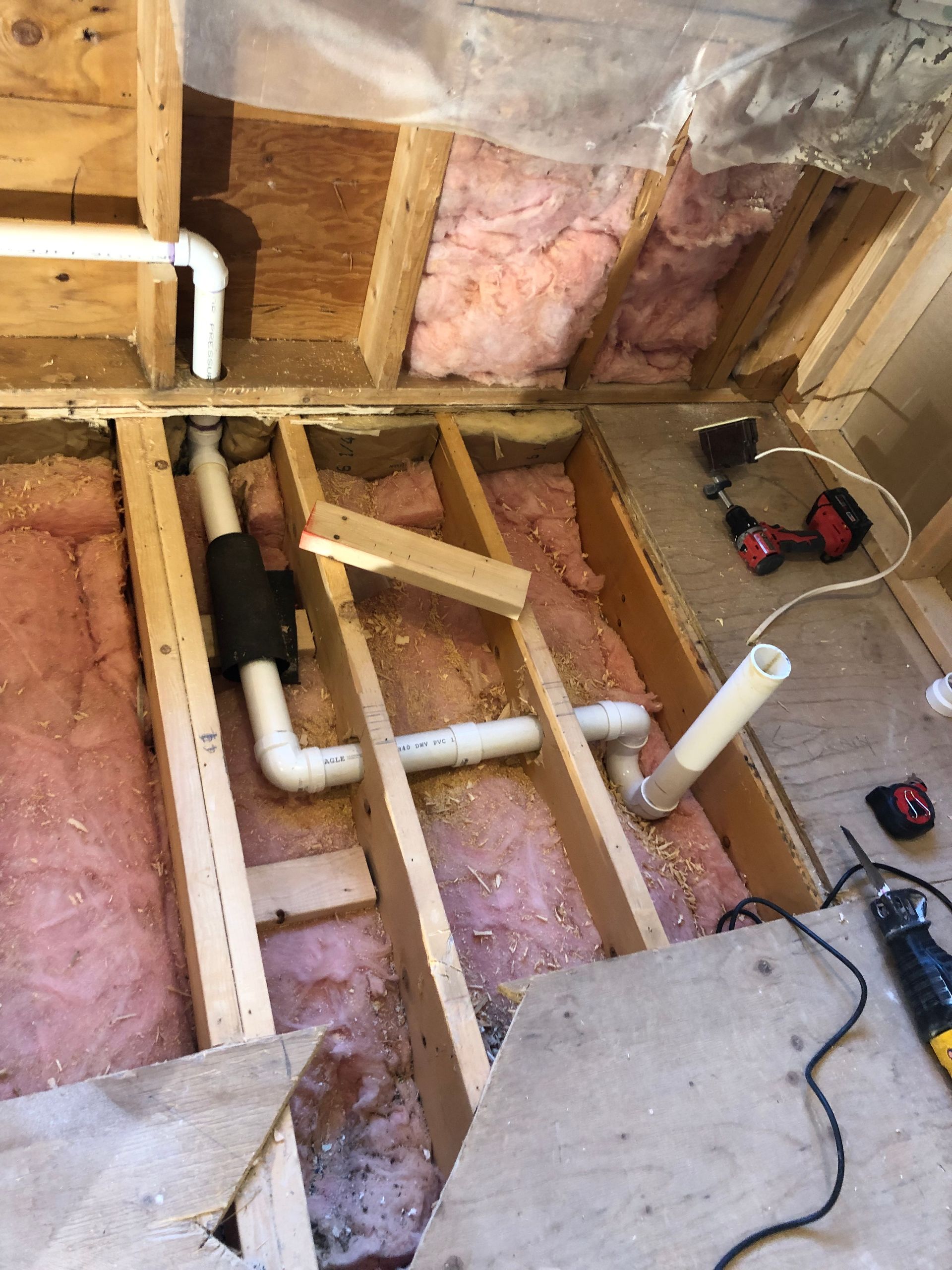 Exposed bathroom floor framing with pink insulation, white PVC pipes, and a cordless drill on the subfloor.