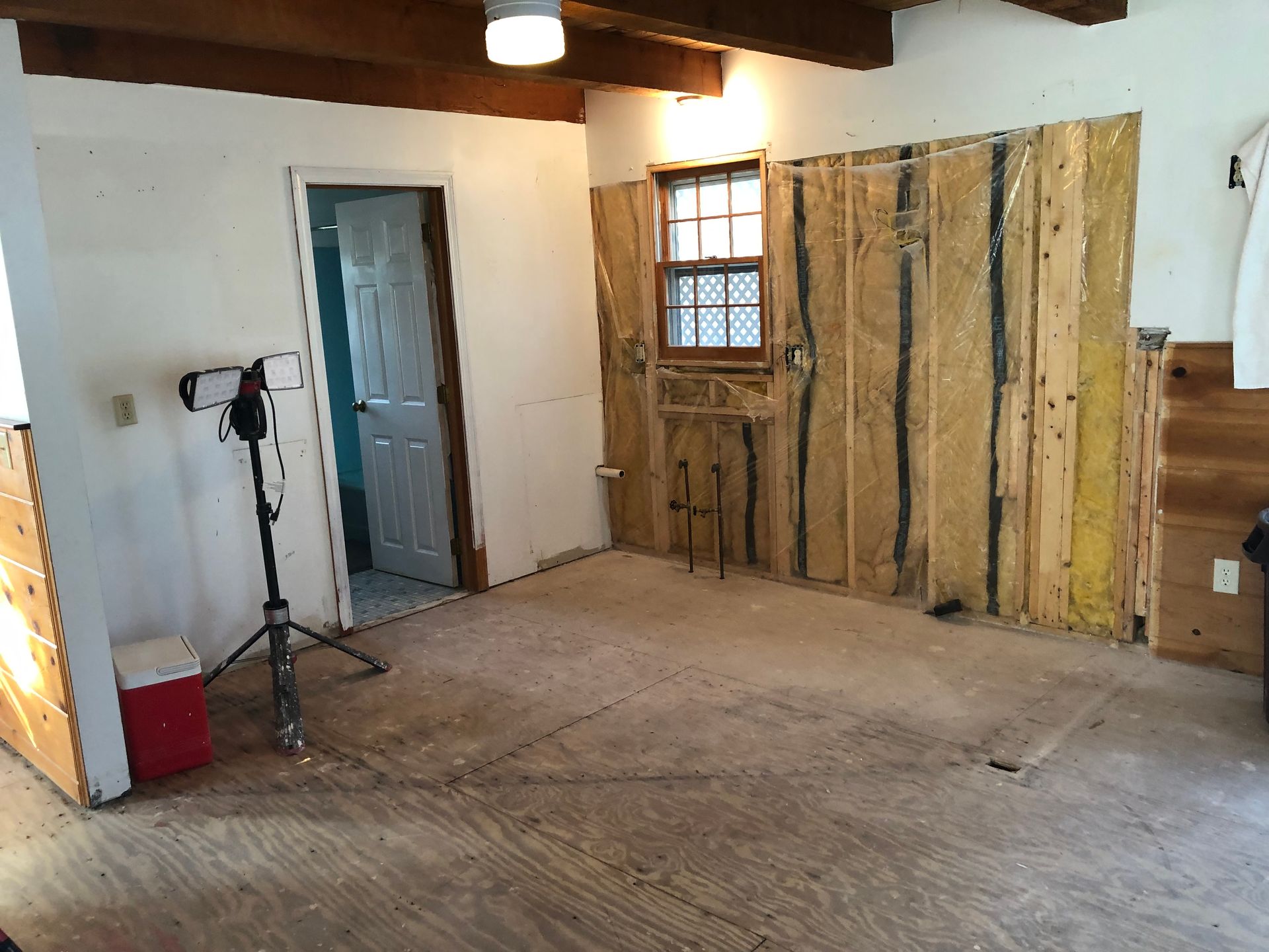 Empty unfinished room with exposed insulation, a door, window, and a tripod light stand on a bare floor.