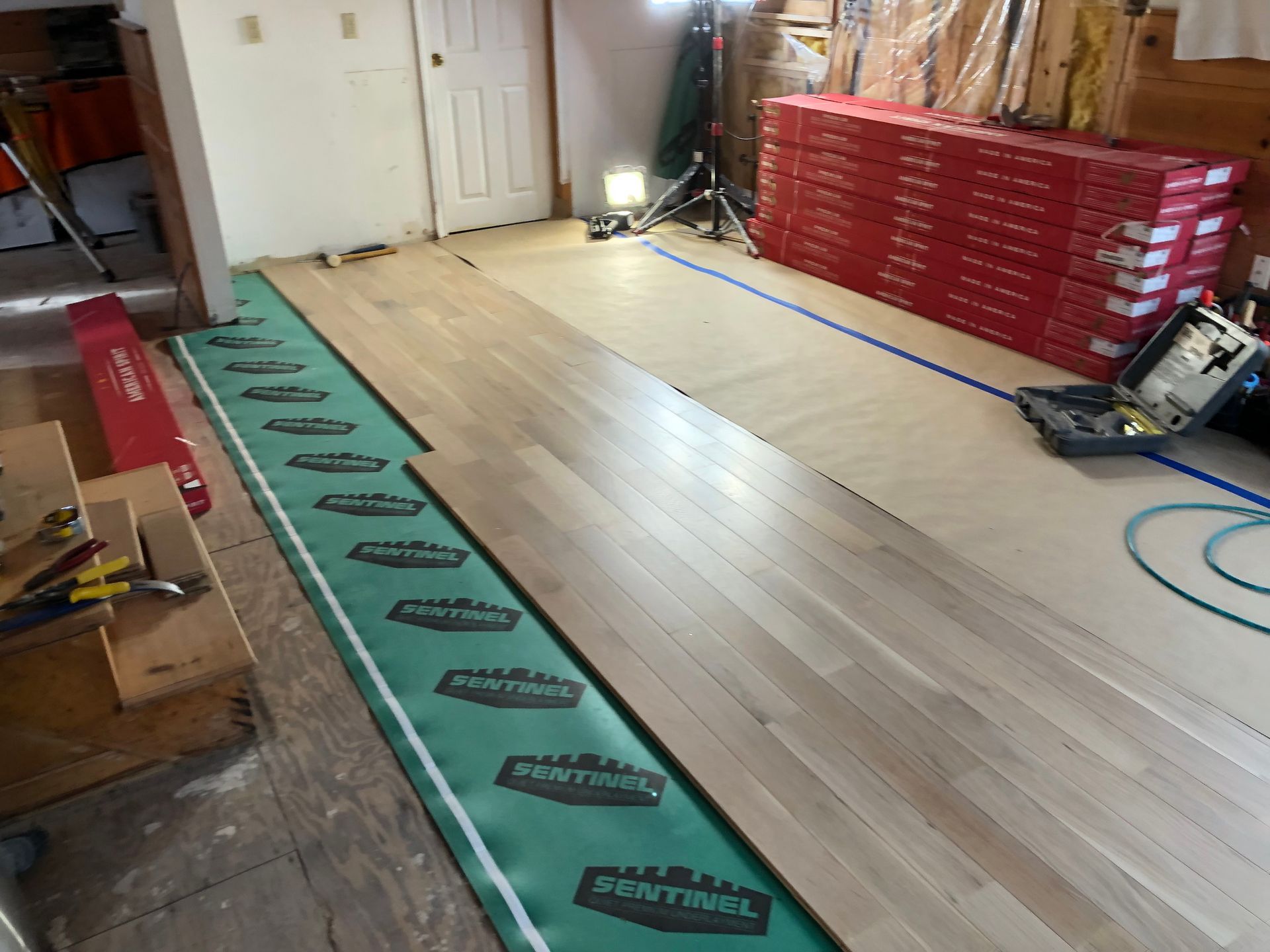 Wood floor installation in progress with green underlayment, red tool chest, and power saw in workshop