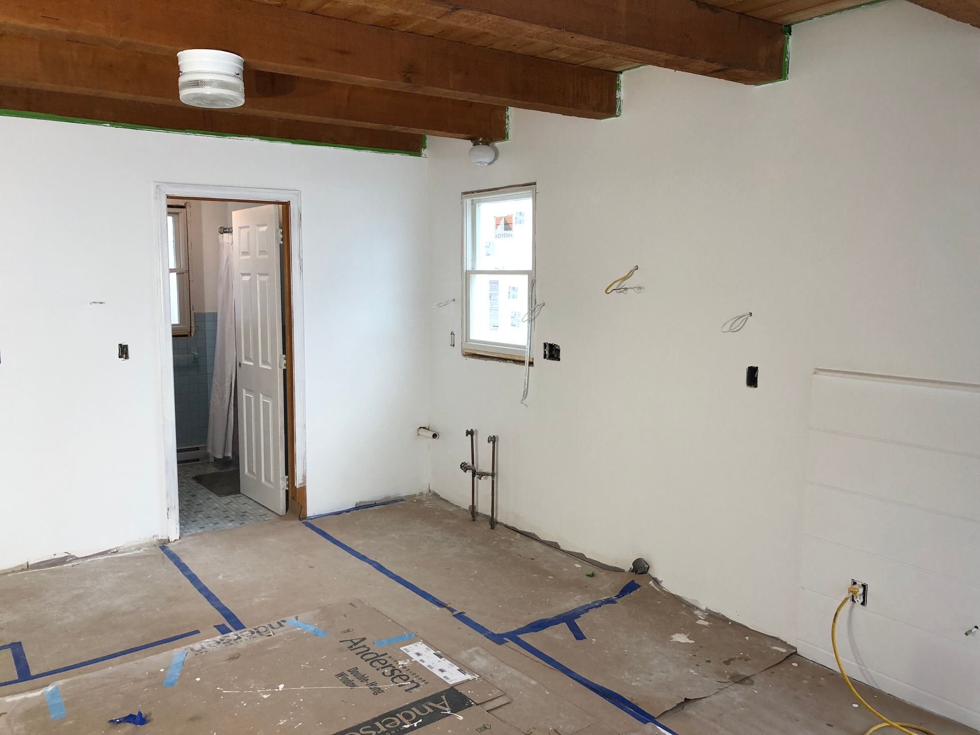 Empty room under renovation with white walls, exposed ceiling beams, and taped-off floor area