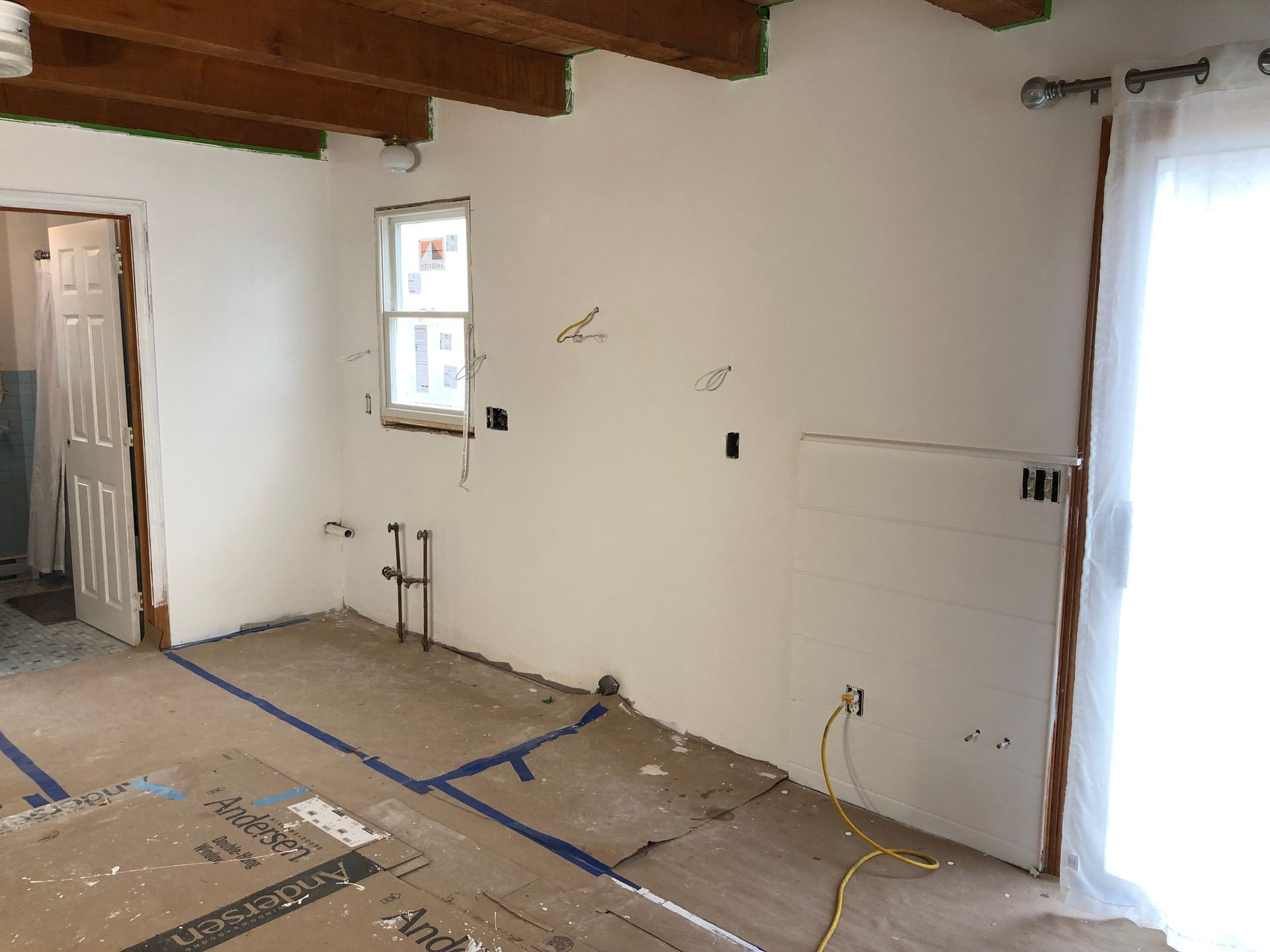Empty room under renovation with exposed beams, taped floor, and bright windows and door on the right.