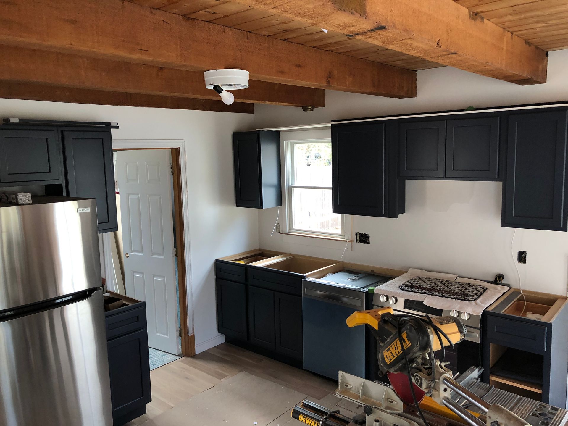 Small kitchen with black cabinets, stainless fridge, wood beams, and a stove on the right.